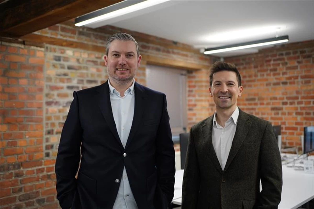 Two people wearing smart jackets and collared shirts stand in a modern office with exposed brick walls, wooden beams, and bright overhead lighting. A conference table and chairs are visible behind them.
