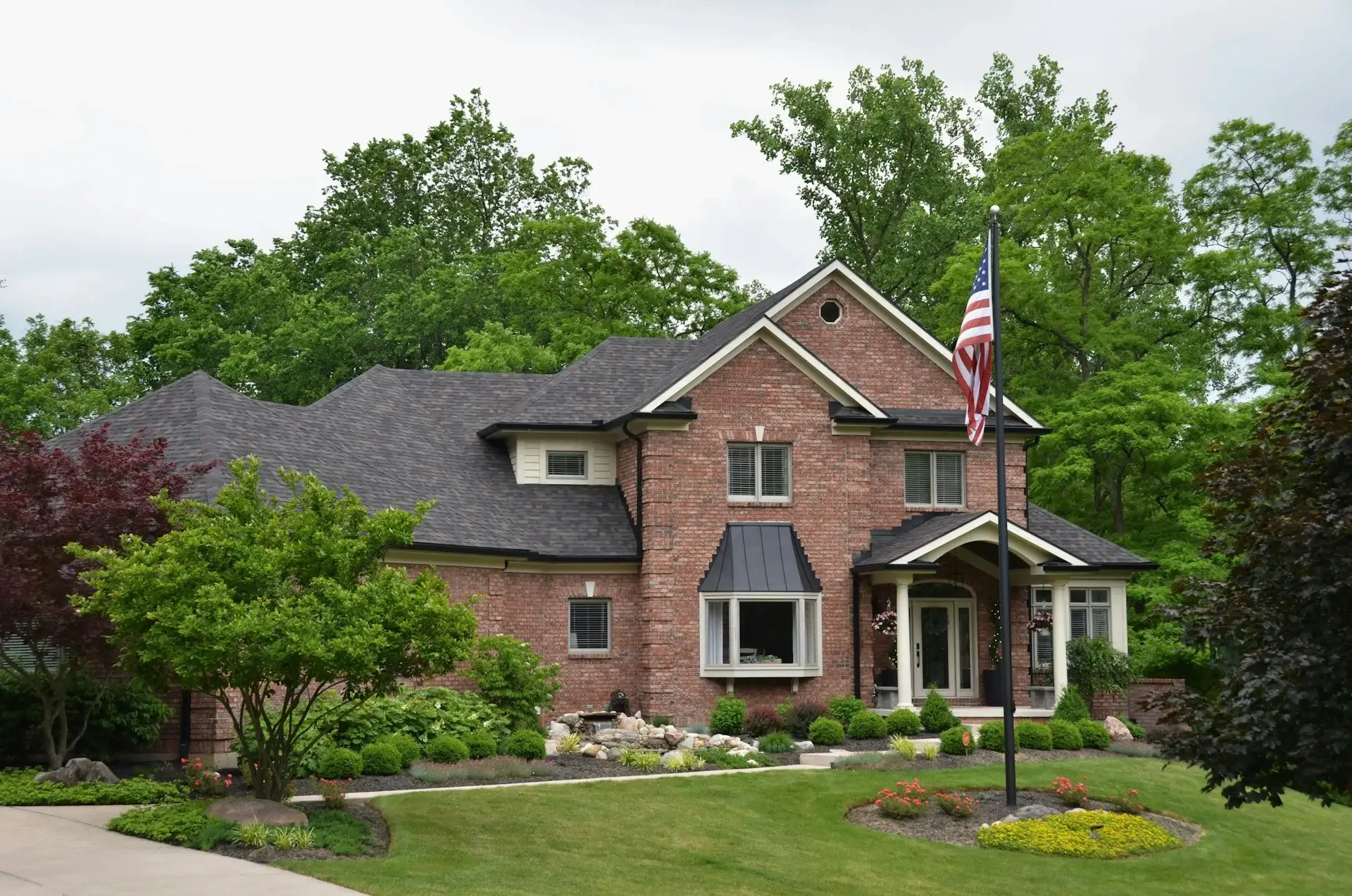 Large red brick house with a dark shingle roof, landscaped garden, front porch entrance, and an American flag in a quiet suburban setting.