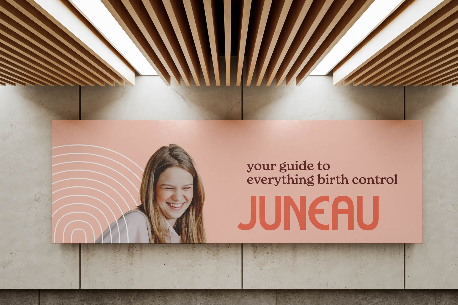 A rectangular sign with a peach-colored background displays a photograph of a smiling woman with long brown hair. White curved lines are visible behind her. Text on the sign reads “your guide to everything birth control” above the word “JUNEAU” in bold, orange letters. The sign is mounted on a light-colored wall with visible concrete or stone panels beneath. Above the sign, a slatted wooden ceiling with recessed lighting is visible.