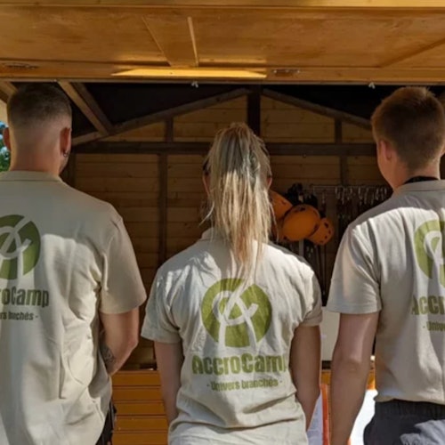 Three people with "AccroCamp" logos on their shirts stand facing away in front of a wooden structure with helmets inside.