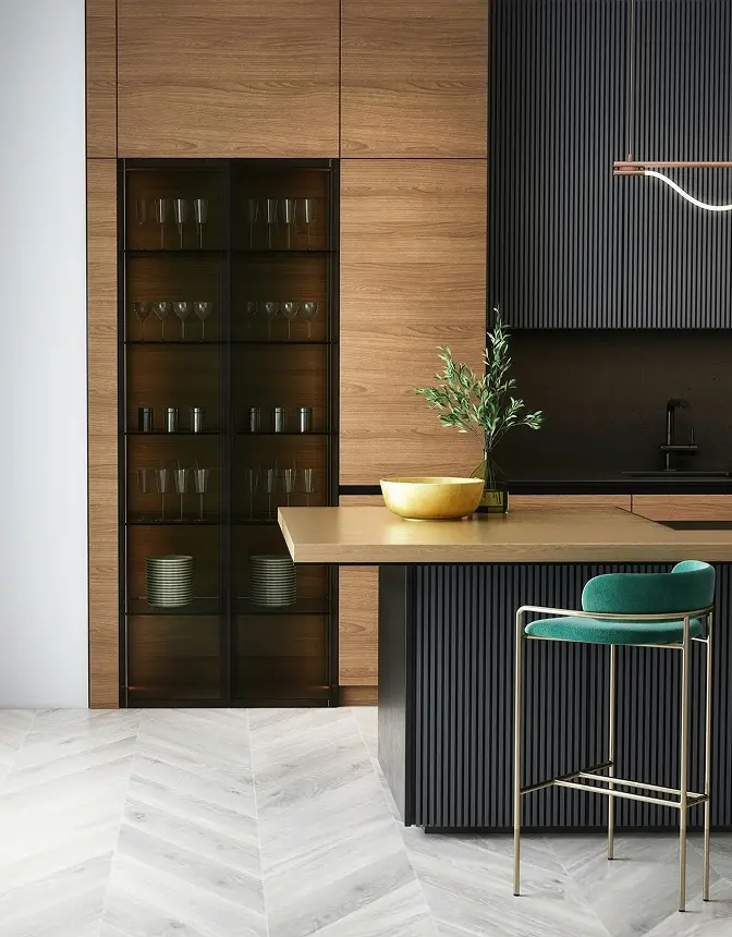 Modern kitchen with wood cabinets, glassware display, and dark backsplash. Features a sleek island, teal bar stool, and decorative bowl with greenery.