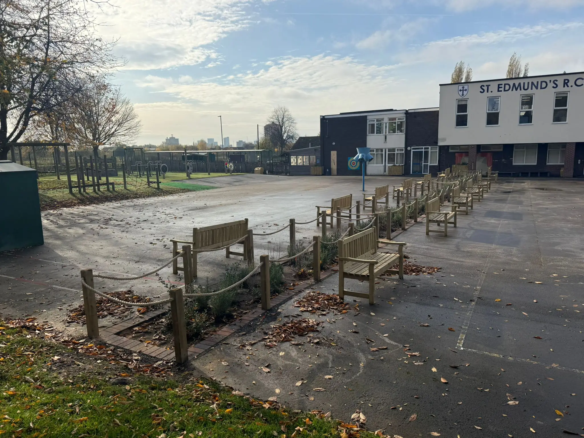An empty lot with overgrown weeds, bordered by a paved walkway and a building in the background under a cloudy sky.