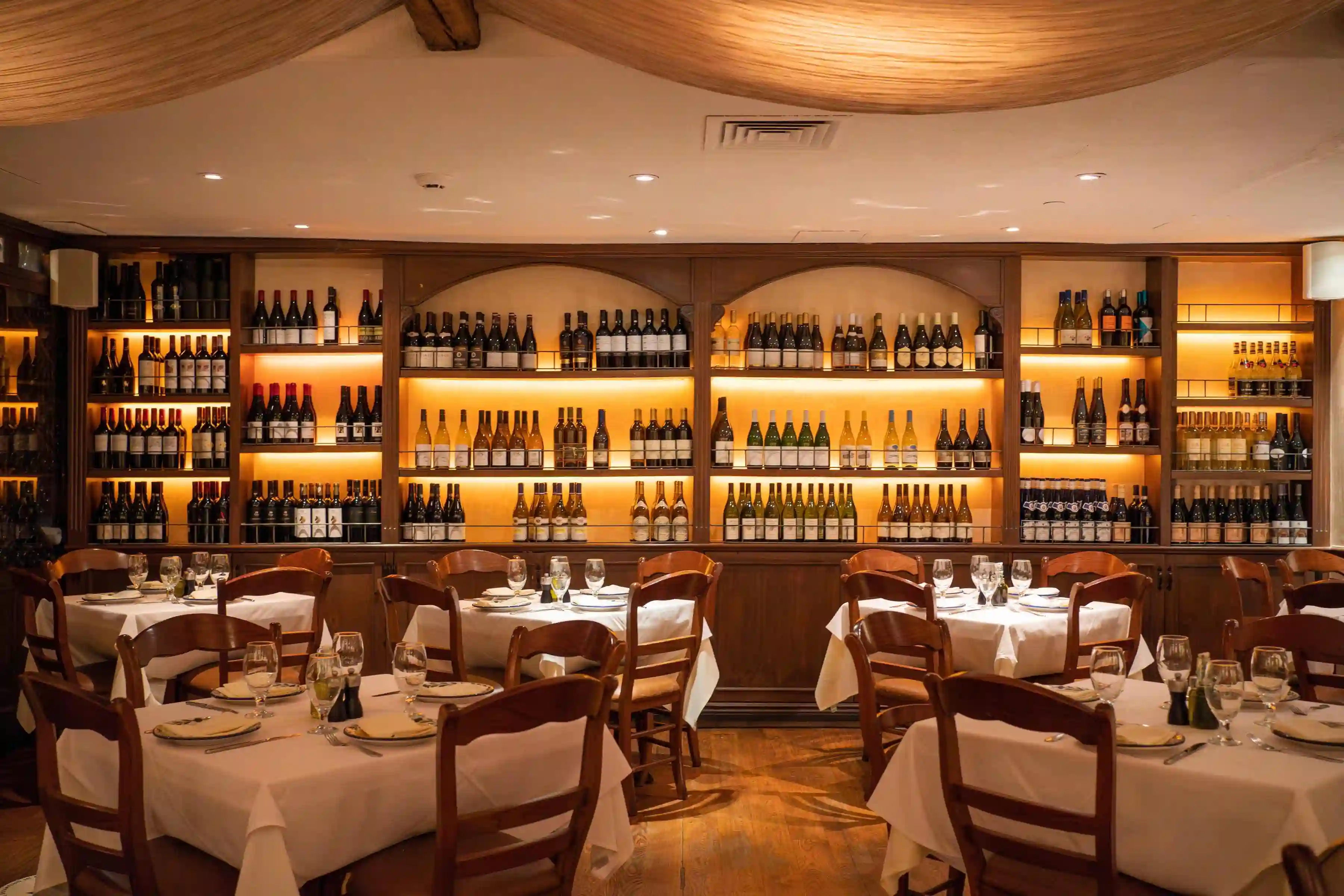 Interior of a restaurant featuring elegant tables, white linens, and a warmly lit shelf display in the background.