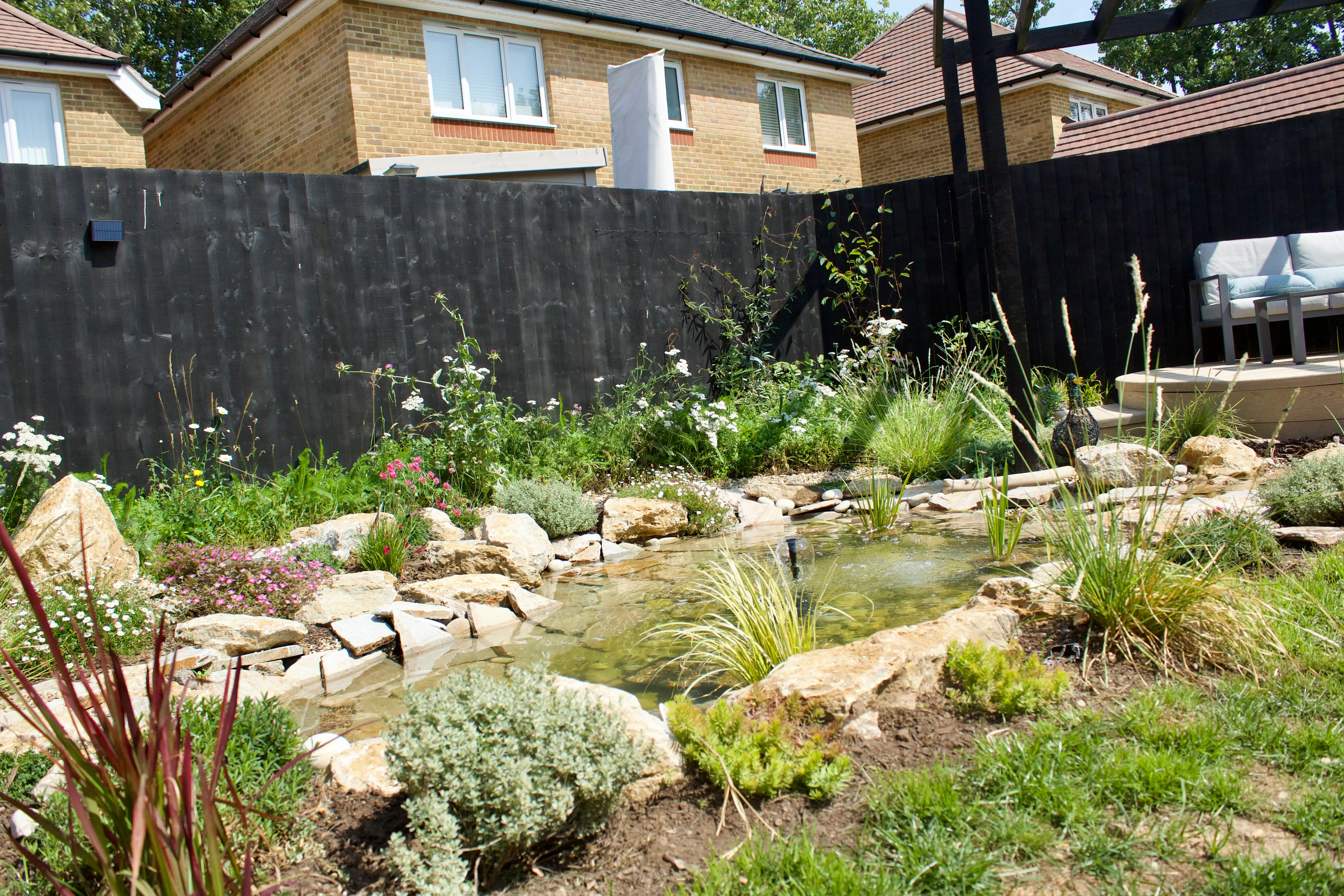 A garden with various plants and rocks, surrounded by a wooden fence and a house in the background.