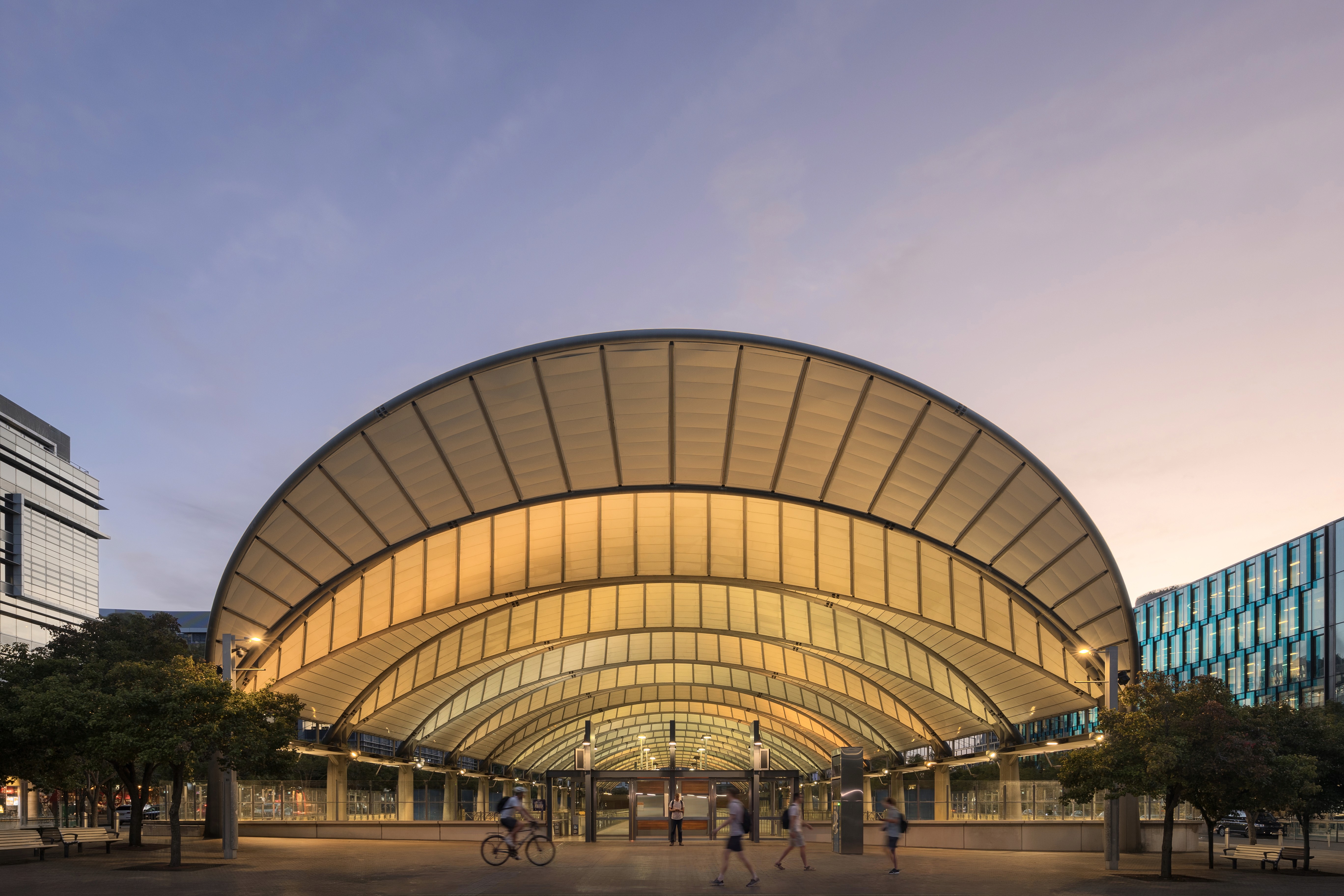 Sydney Olympic Park Train Station at dusk, where its arched roof and structural rhythm define a high-performance transport hub, captured in motion to reveal how contemporary transport architecture supports real-world circulation and spatial performance.