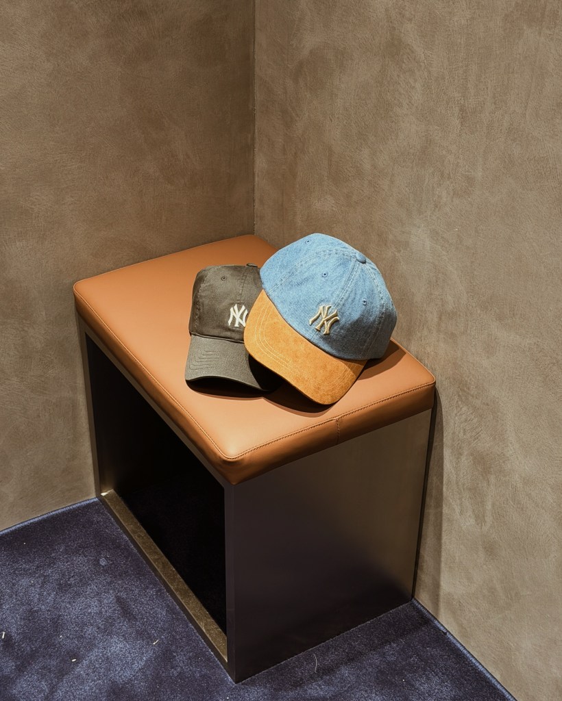 Two baseball caps with the New York logo, one in olive green and the other in denim blue with an orange peak, resting on a stylish bench in a contemporary store setting.