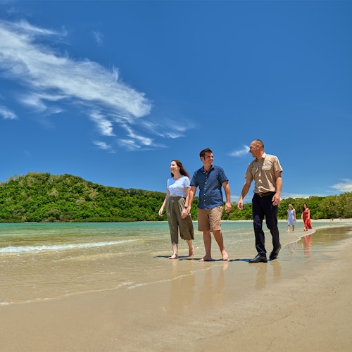 Tre persone camminano lungo una spiaggia di sabbia vicino a un'acqua limpida, con colline verdi e un cielo azzurro con alcune nuvole sullo sfondo.
