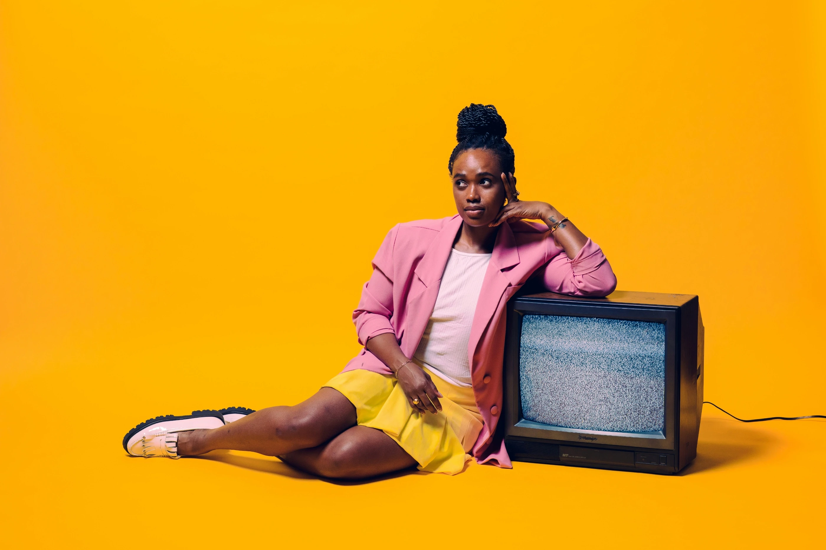 Frau sitzt nachdenklich vor gelbem Hintergrund und lehnt an einem alten Fernseher mit statischem Bild, farblich kontrastierende Kleidung, inszenierte Studioaufnahme.