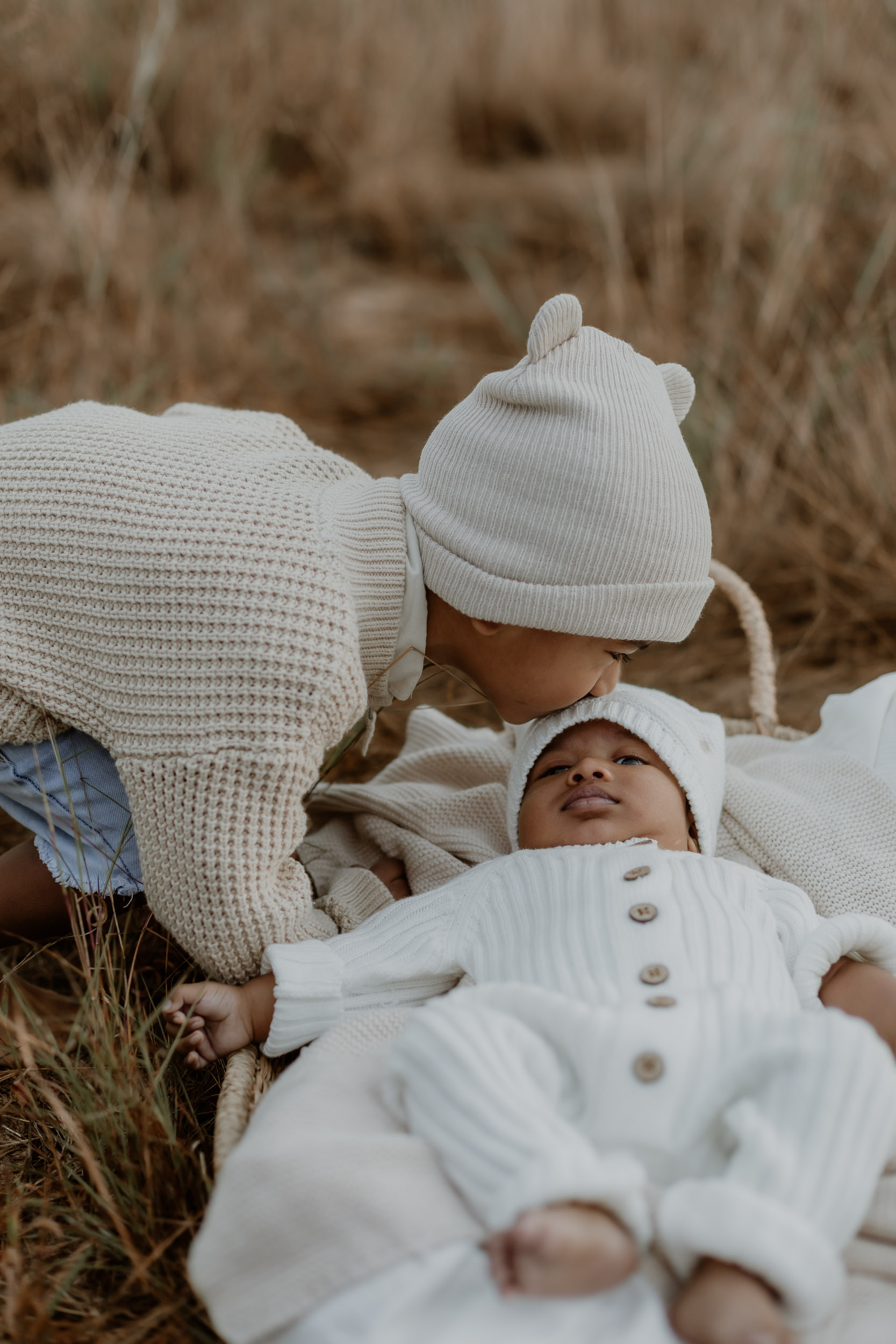 Sibling kissing newborn baby in grassy outdoor newborn session