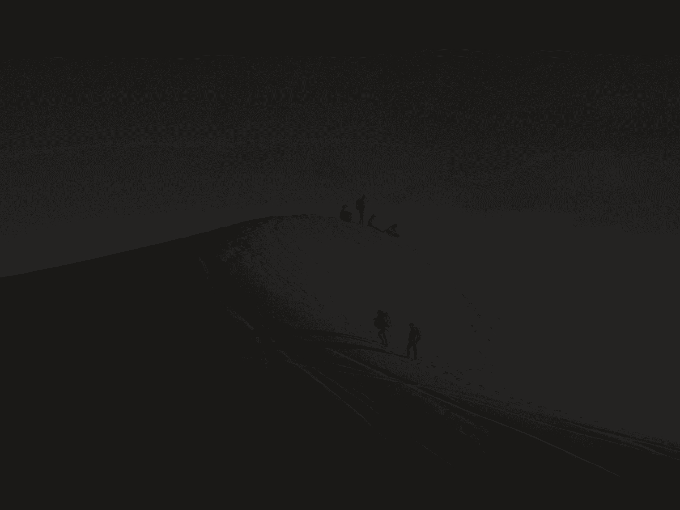Silhouetted figures hiking and resting on a dark sand dune ridge.