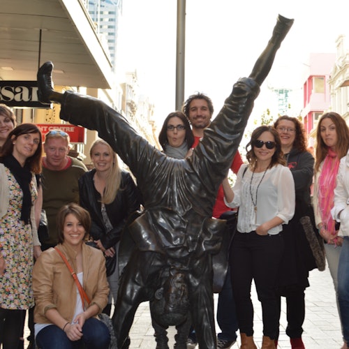 Group of people posing around an upside-down statue on a city street, with buildings and shop signs in the background.
