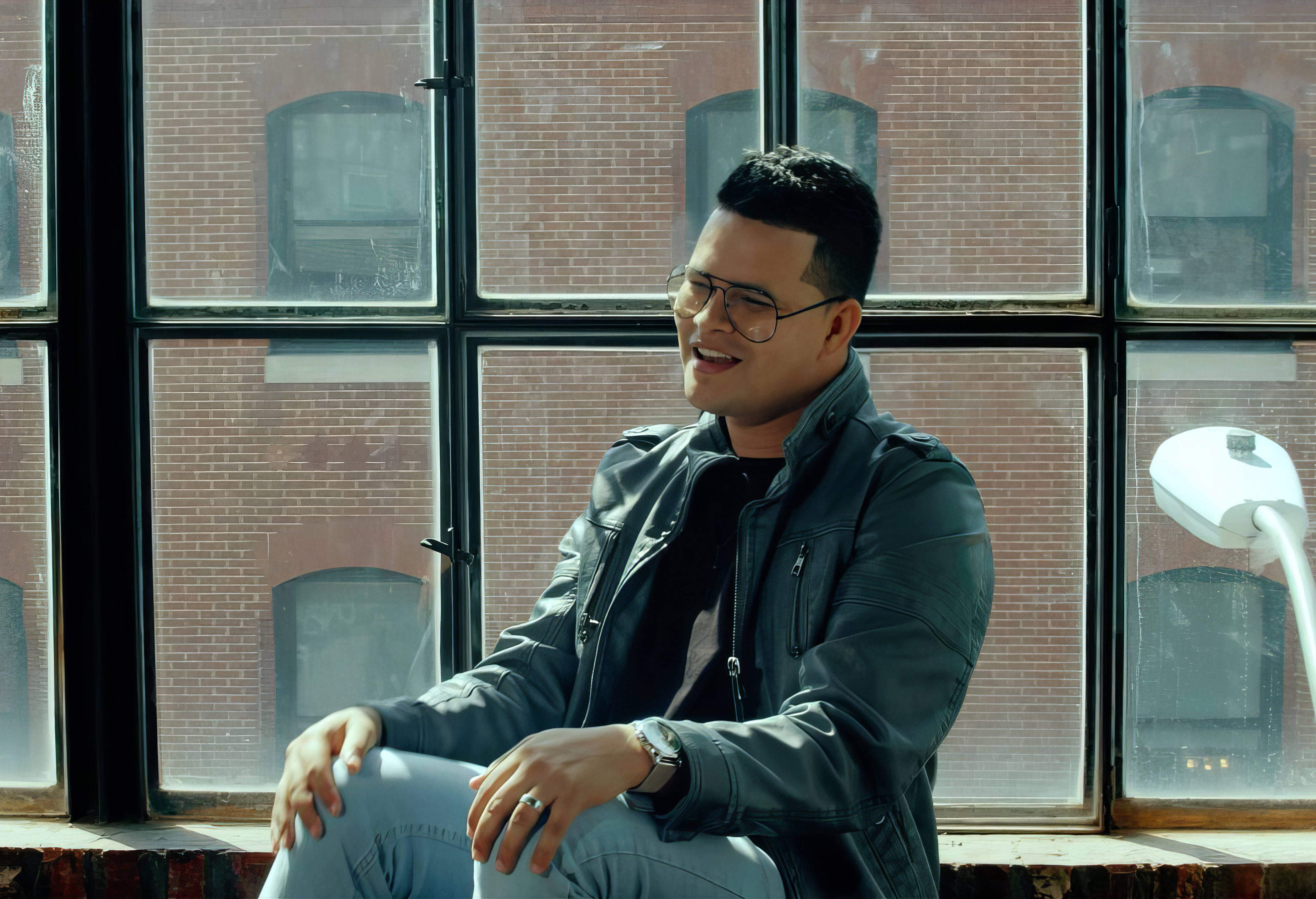 Dani Sierra sitting by a large window, smiling, wearing a denim jacket and casual attire.