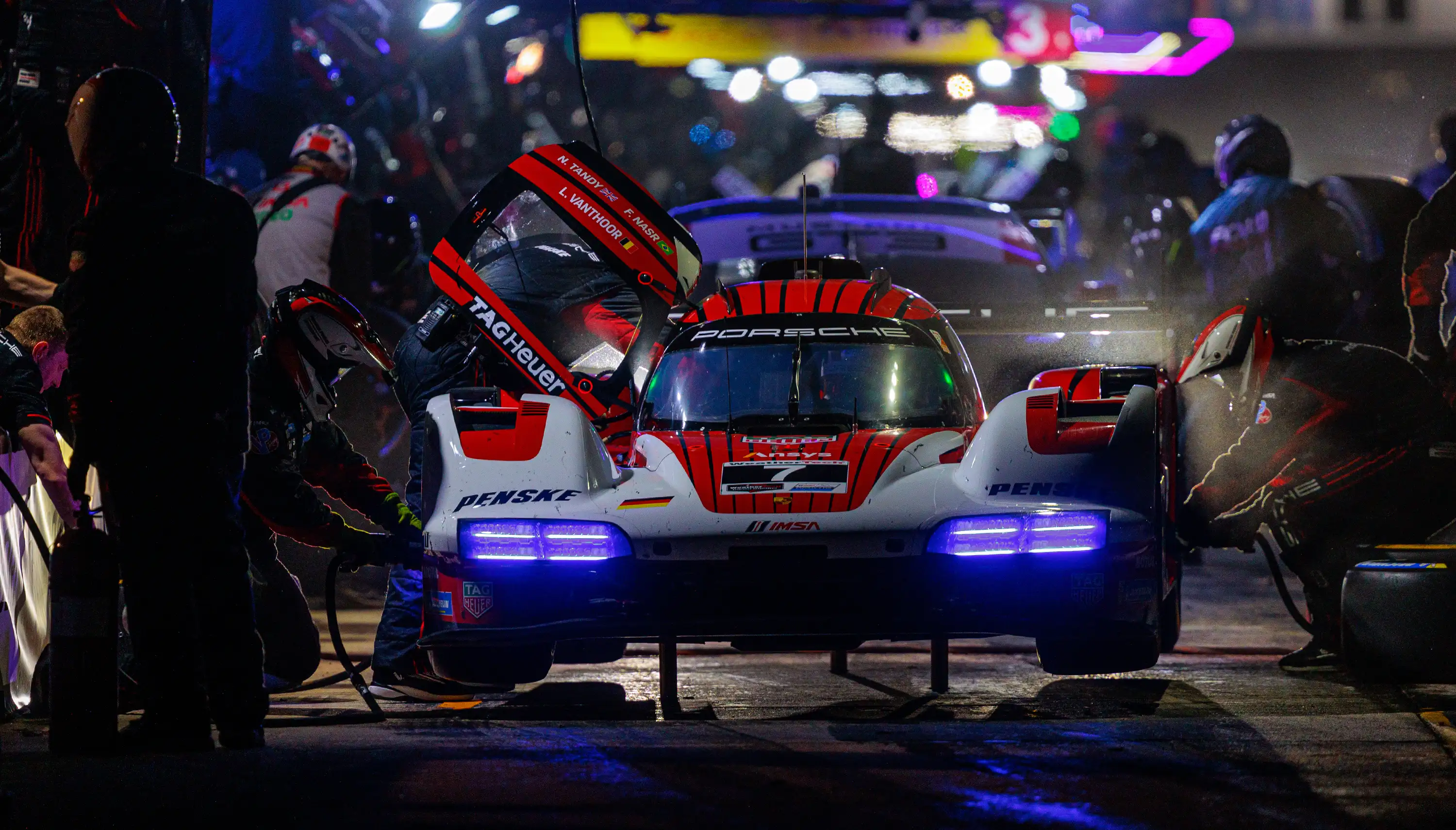 A mechanic repairs a red and white Porsche race car in a pit stop, focusing on the engine while tools are scattered around the workspace.