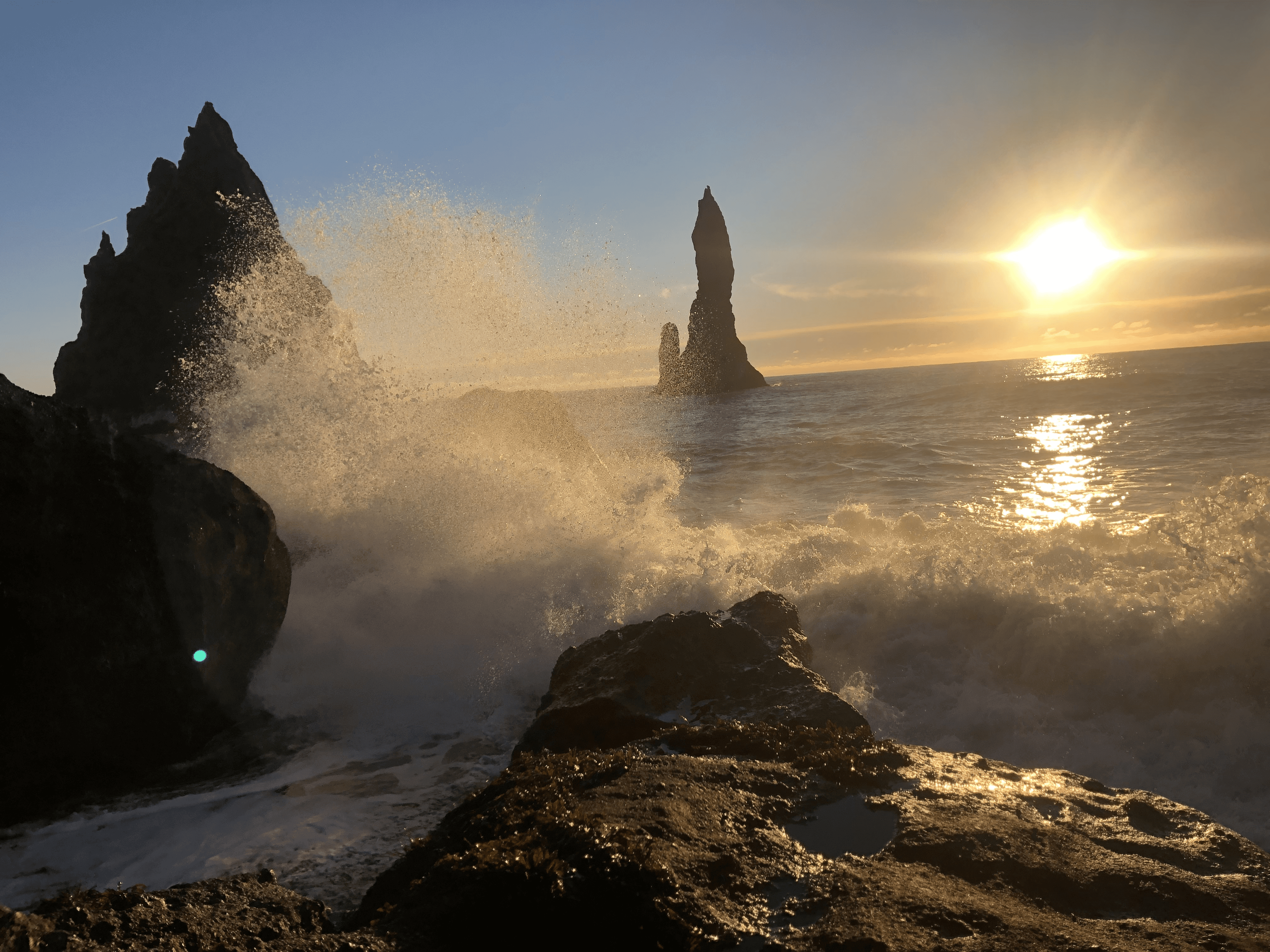 Dramatic ocean waves crash against black rock spires at Reynisfjara beach under golden light, evoking a romantic and powerful atmosphere.