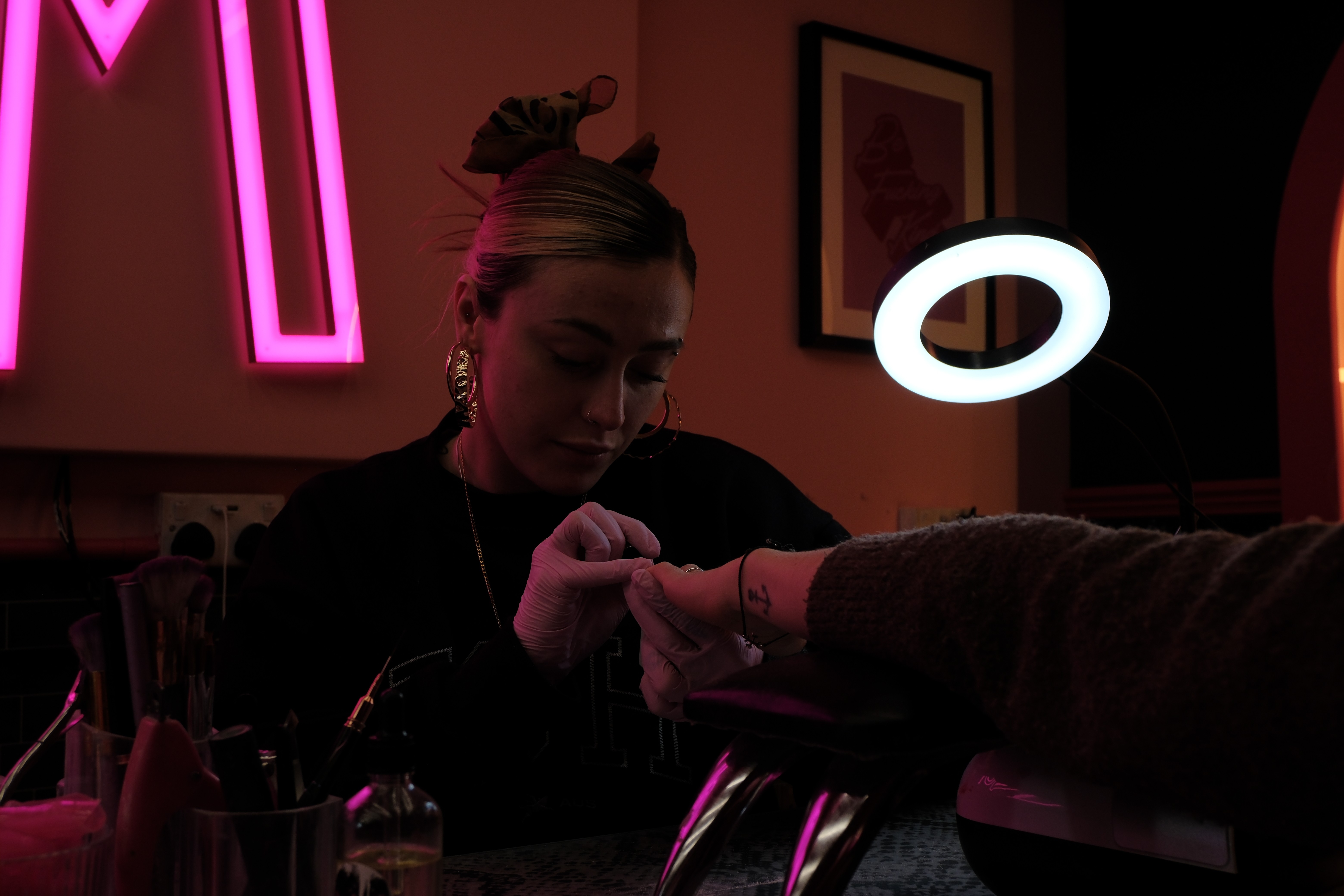A medium close-up of a technician with a leopard-print hair bow and large gold hoops. She is wearing pink nitrile gloves and looking down with deep concentration as she works on a client's hand. A bright ring light illuminates her workspace against the dim, pink-hued background.