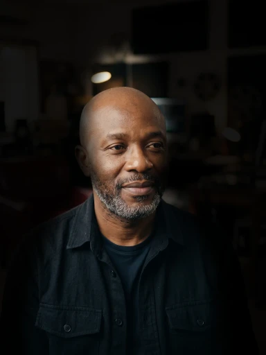 Jude Mensah, grey beard and a black shirt, in a dim studio