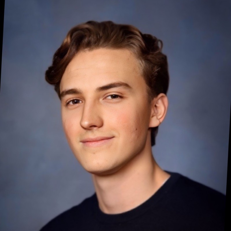 A focused woman with long red hair is seated at a desk, wearing a black blazer and dark blouse. She intently looks at camera, smiling slightly, with an open laptop in front of her. The setting is a warm and serene office setup in the background. Soft daylight filters through, creating a calm atmosphere. The composition captures her in a frontal profile with a clear emphasis on her concentration and the serene indoor setting.