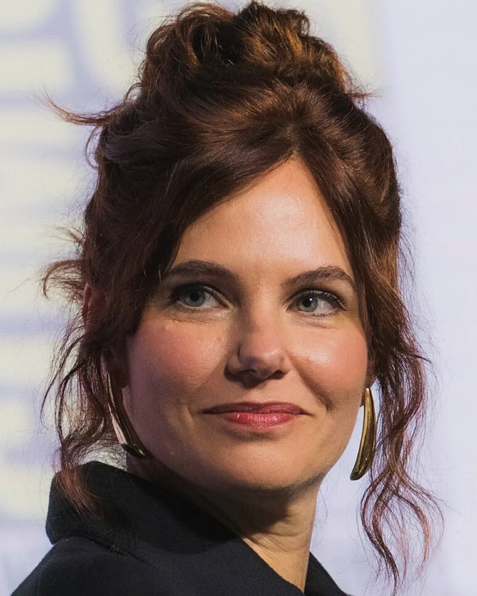 A woman with auburn hair styled in a loose updo and wearing gold hoop earrings is looking slightly to the side against a softly blurred background.