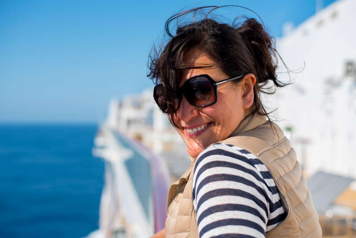 A smiling woman with dark hair in a ponytail and sunglasses smiles while wearing a striped shirt on a cruise ship deck.