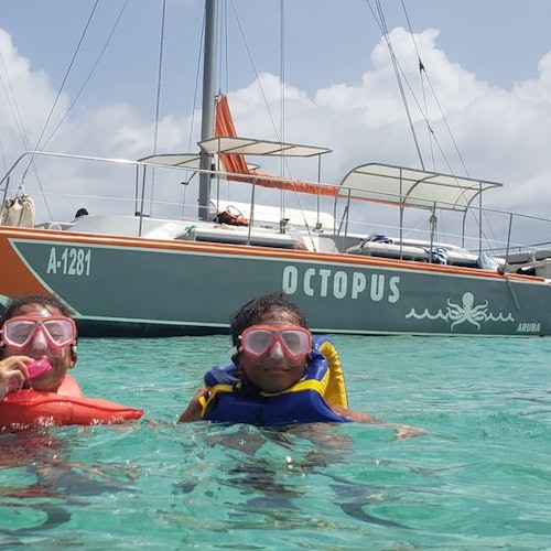 Deux enfants portant des équipements de plongée avec tuba et des gilets de sauvetage nagent dans une eau turquoise claire près d'un voilier nommé "Octopus."