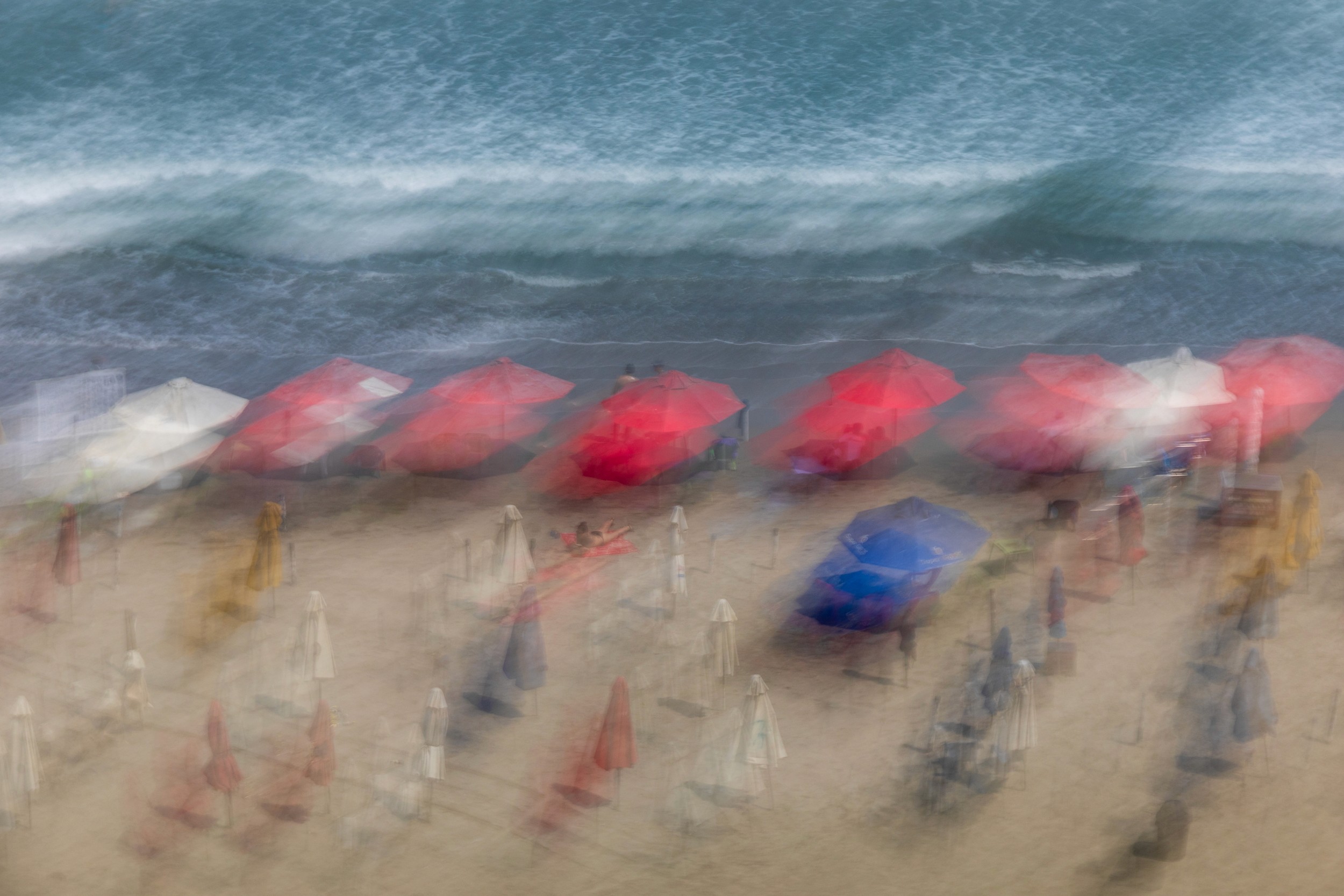 Red umbrellas and beachgoers appear painterly and scattered against a textured ocean backdrop