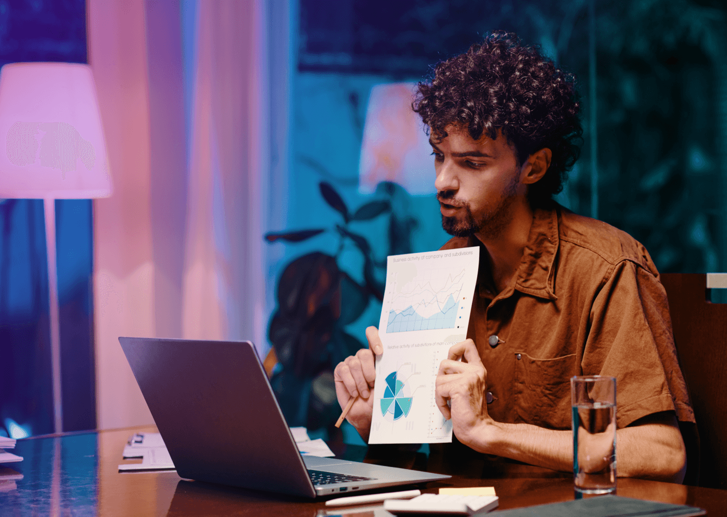 Digital marketing specialist presenting data and analytics during an online meeting, showcasing business growth charts on paper, with a laptop on the desk in a professional yet relaxed home office setting