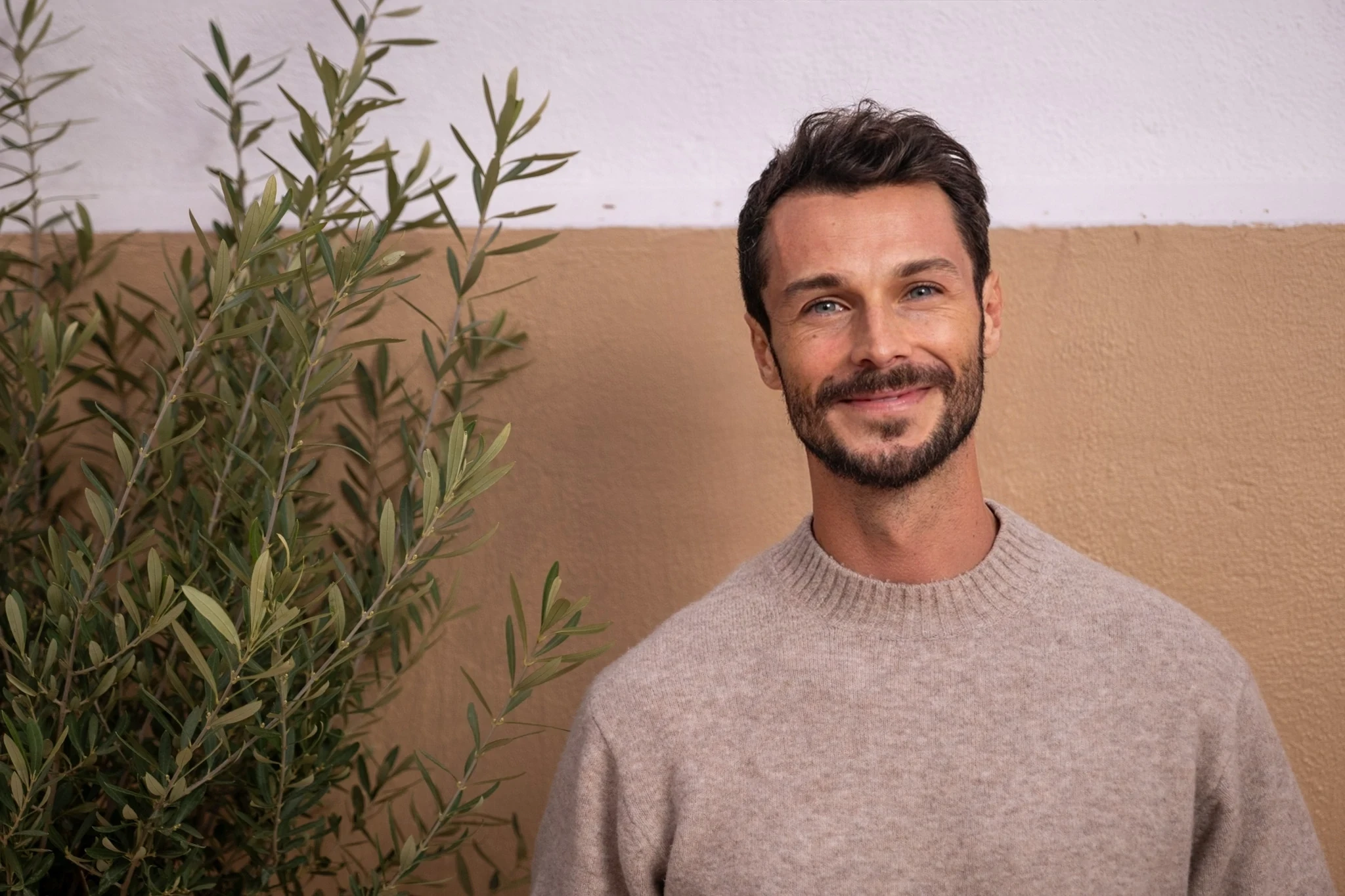 Portrait souriant d'un homme barbu devant un fond clair et une plante. Look décontracté.