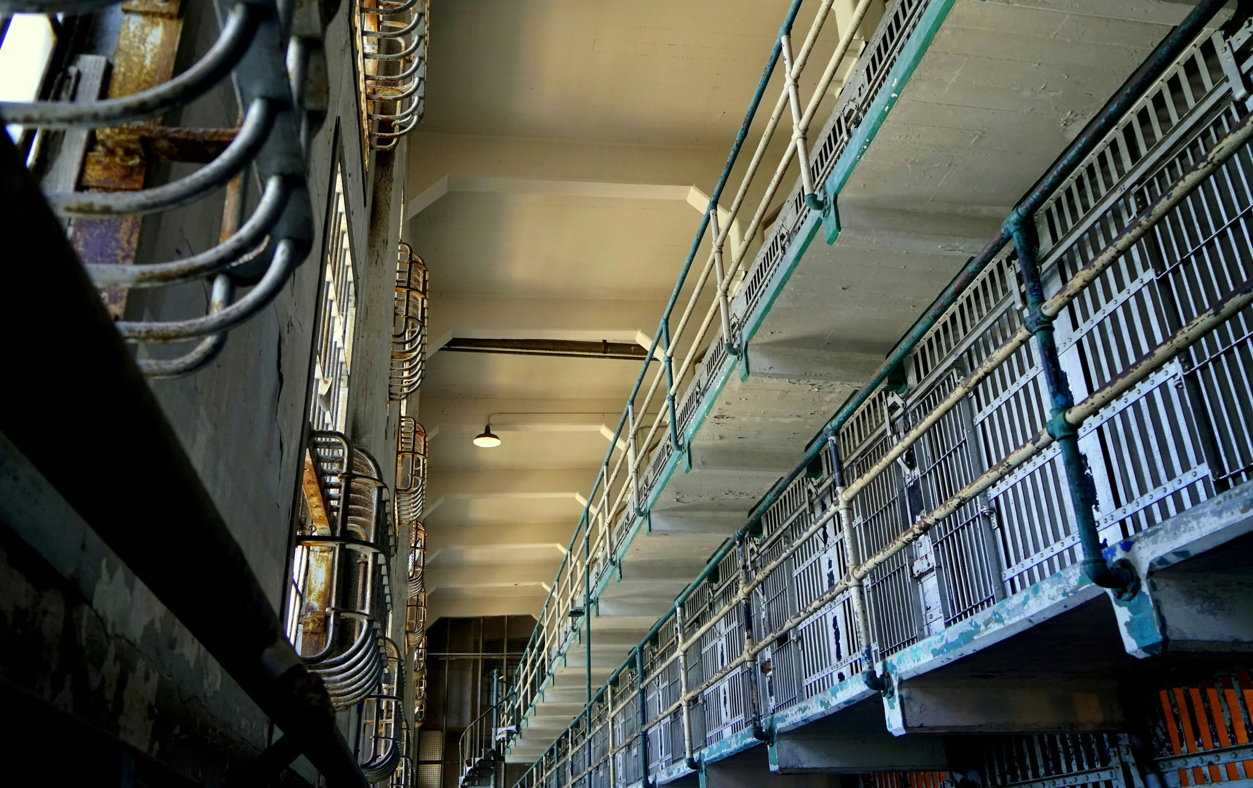 A imagem mostra o interior de um antigo bloco de celas de prisão com vários níveis, com corrimãos e varandas de metal, capturando a arquitetura austera e industrial e a iluminação fraca típicas de penitenciárias históricas.
