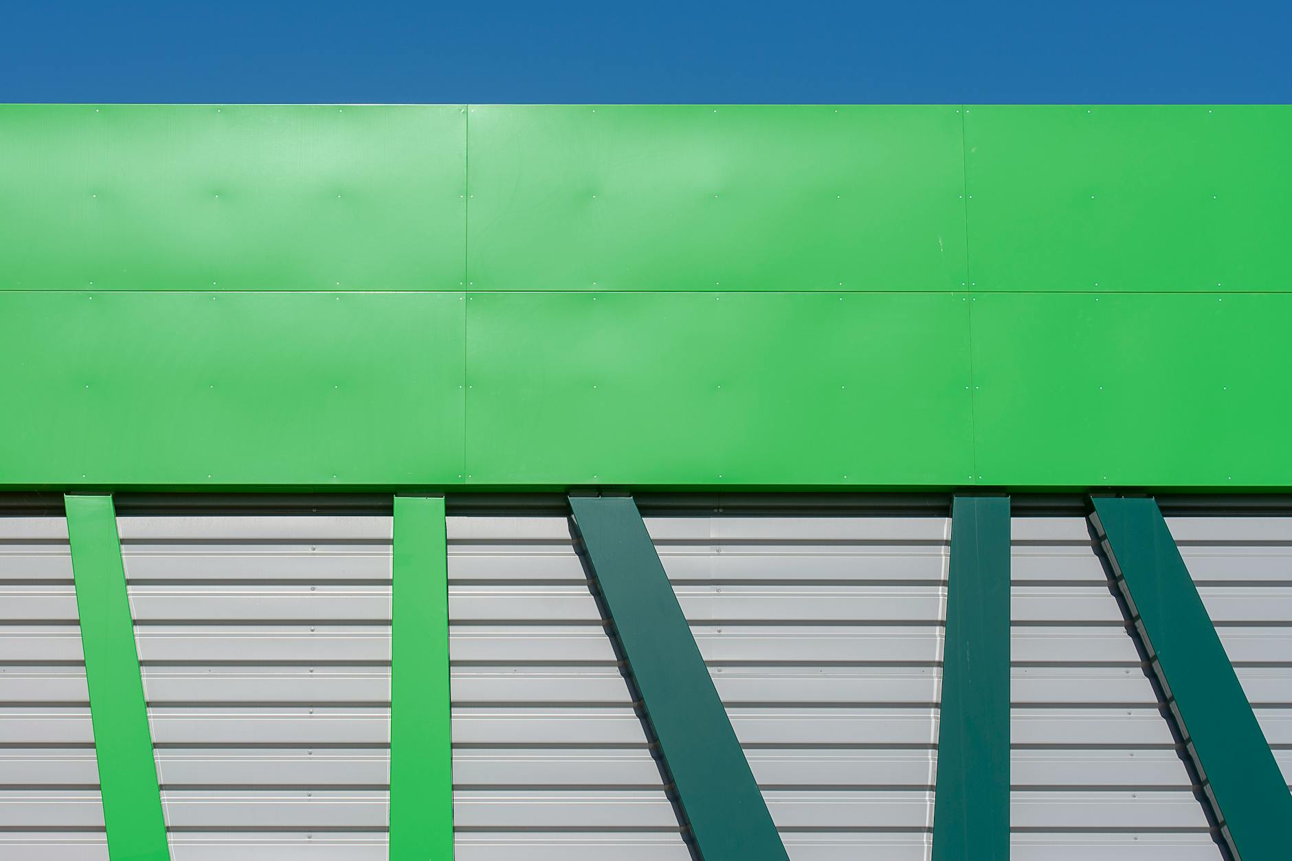 Building exterior with green cladding panels above silver horizontal cladding and green vertical supports against blue sky