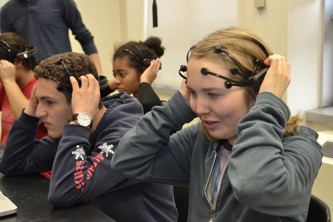 Students in a classroom adjust Emotiv EEG headsets during a neuroscience learning activity. Several students sit at lab tables preparing for a brain-computer interface or cognitive research exercise while an instructor stands nearby. The participants appear focused as they fit the headsets before beginning the session.