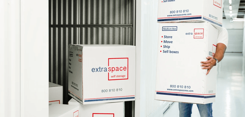 A person carries stacked boxes labeled "Extra Space Self Storage" in a storage facility with an open unit in view. The setting is organised and efficient.