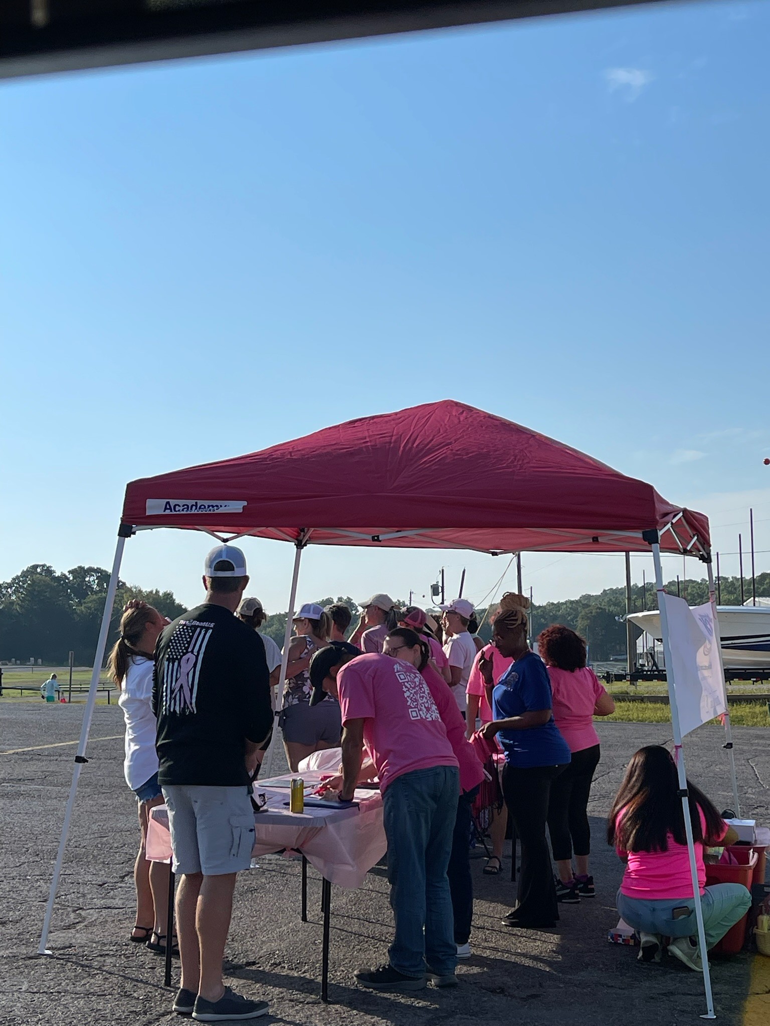 A group of people gather under a red canopy tent on a sunny day, participating in an outdoor event, with some wearing pink shirts as they engage in activities around a table set on a paved surface.