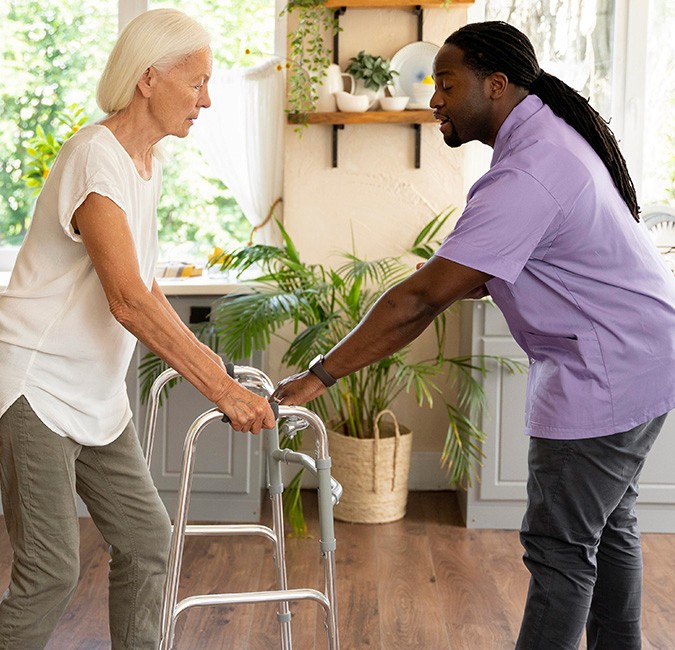 A male caregiver in a purple uniform assisting an elderly woman as she uses a walker in a bright home setting