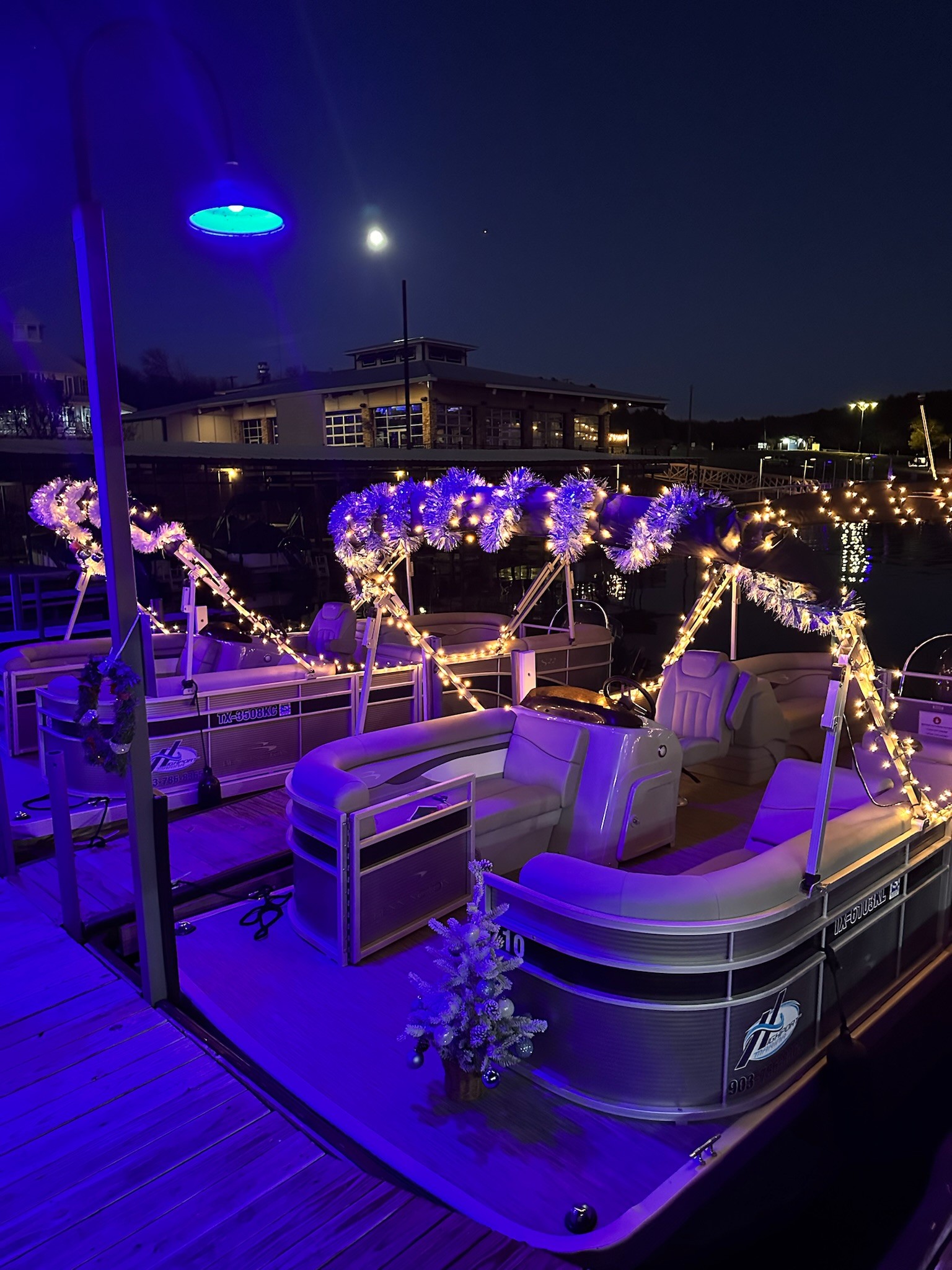 A row of illuminated pontoon boats decorated with string lights and festive garlands are docked at a marina under a clear night sky with a full moon overhead.