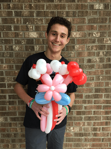Smiling teenage Daniel holding a colorful bouquet of balloon flowers against a brick wall background