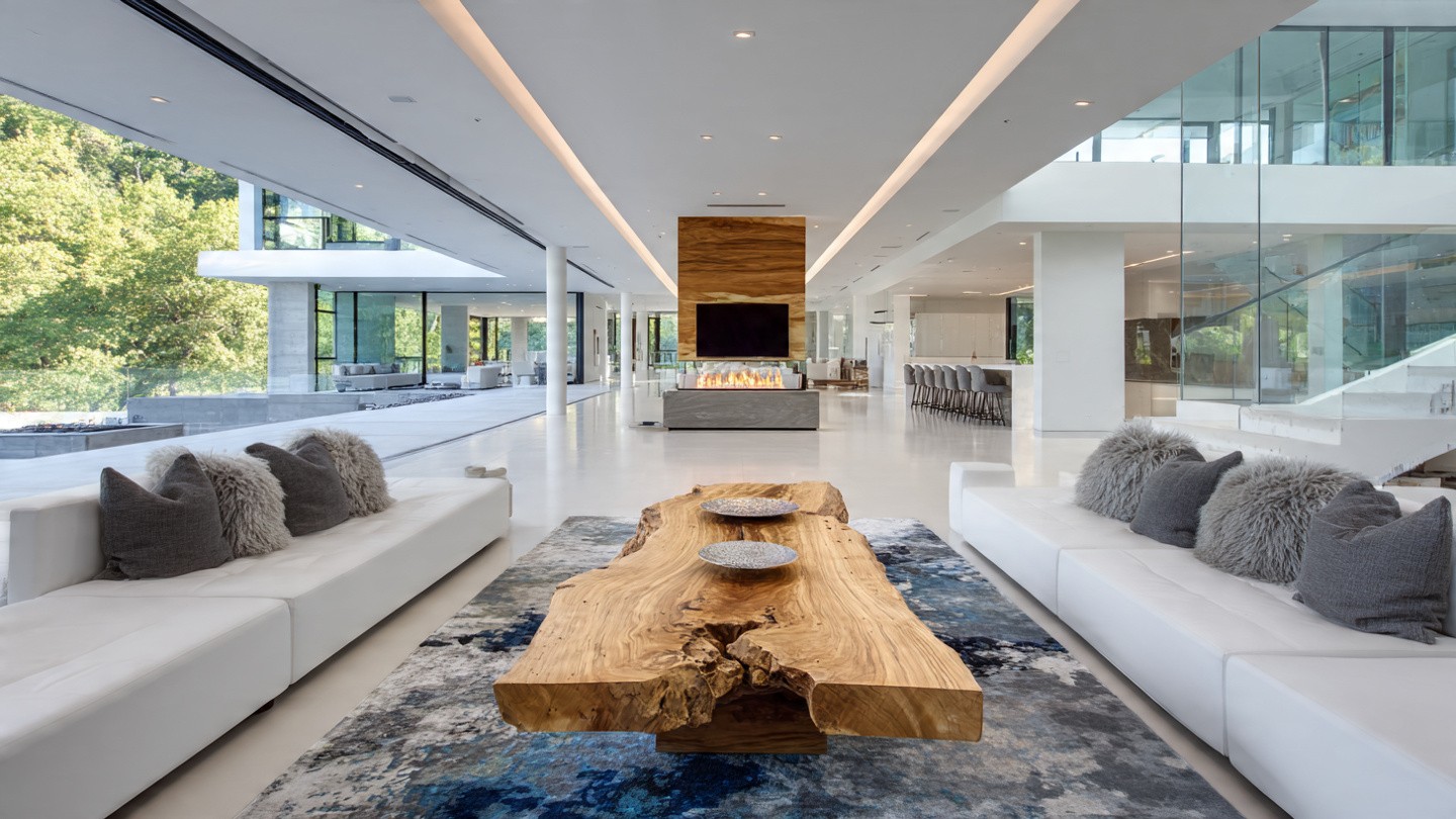 Luxurious modern living room with large glass windows, white sofas, gray cushions, and a natural wood coffee table on a blue-patterned rug. Bright and airy ambiance.