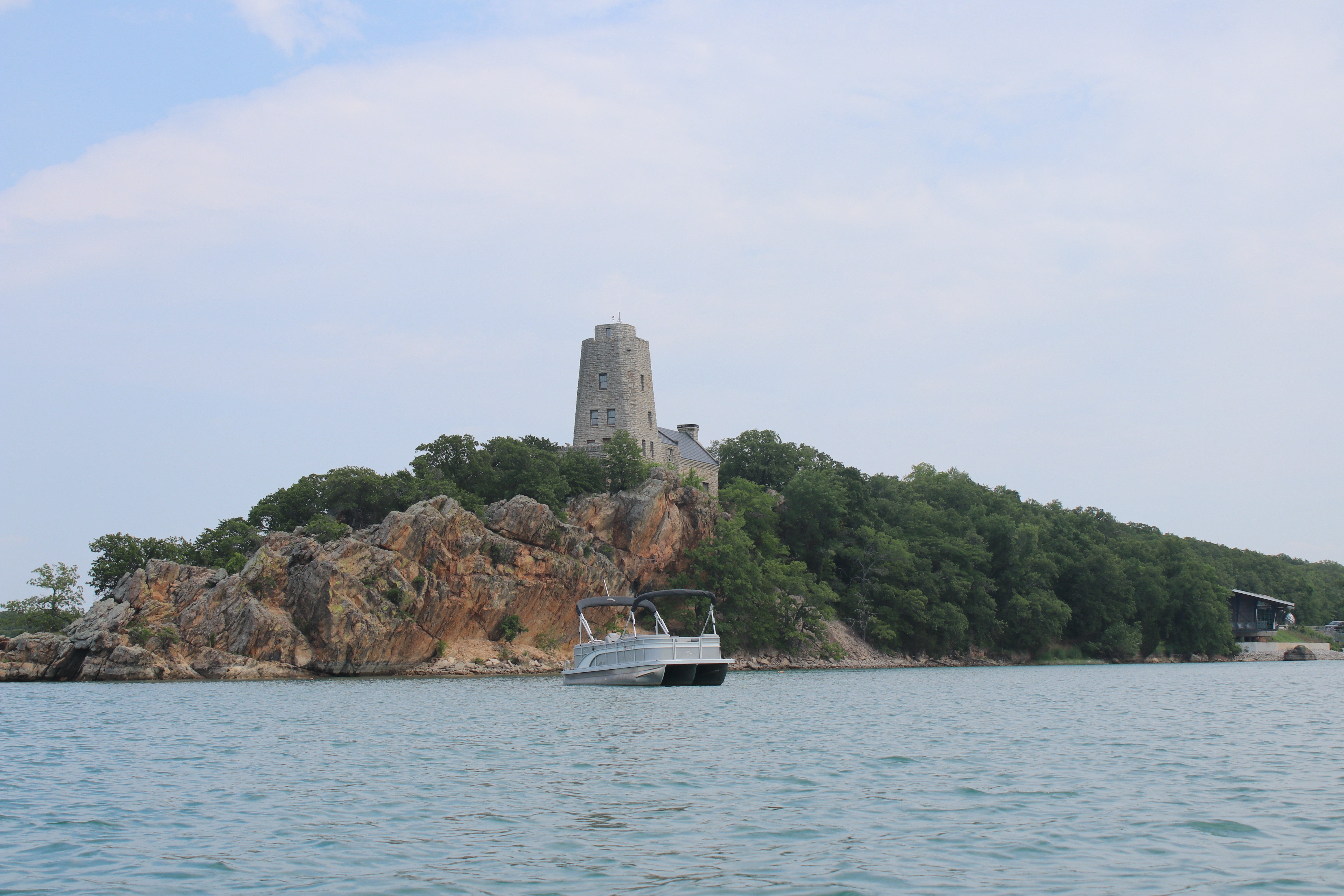 A Tritoon boat is anchored on the tranquil waters near a rocky shoreline, with a historic stone tower surrounded by lush greenery in the background at Lake Murray Marina.