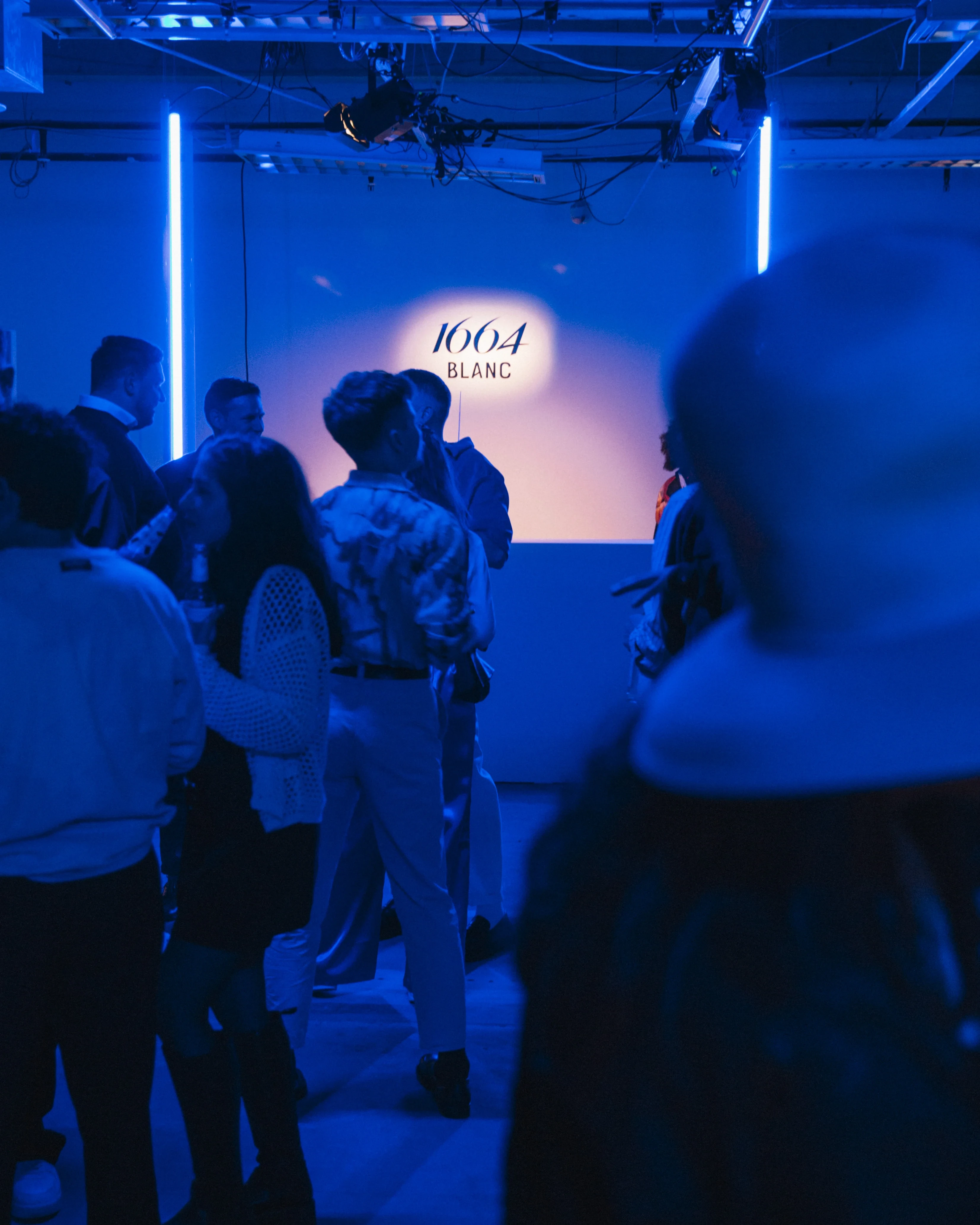 Crowd in blue light in front of the illuminated 1664 Blanc logo wall at the launch event.