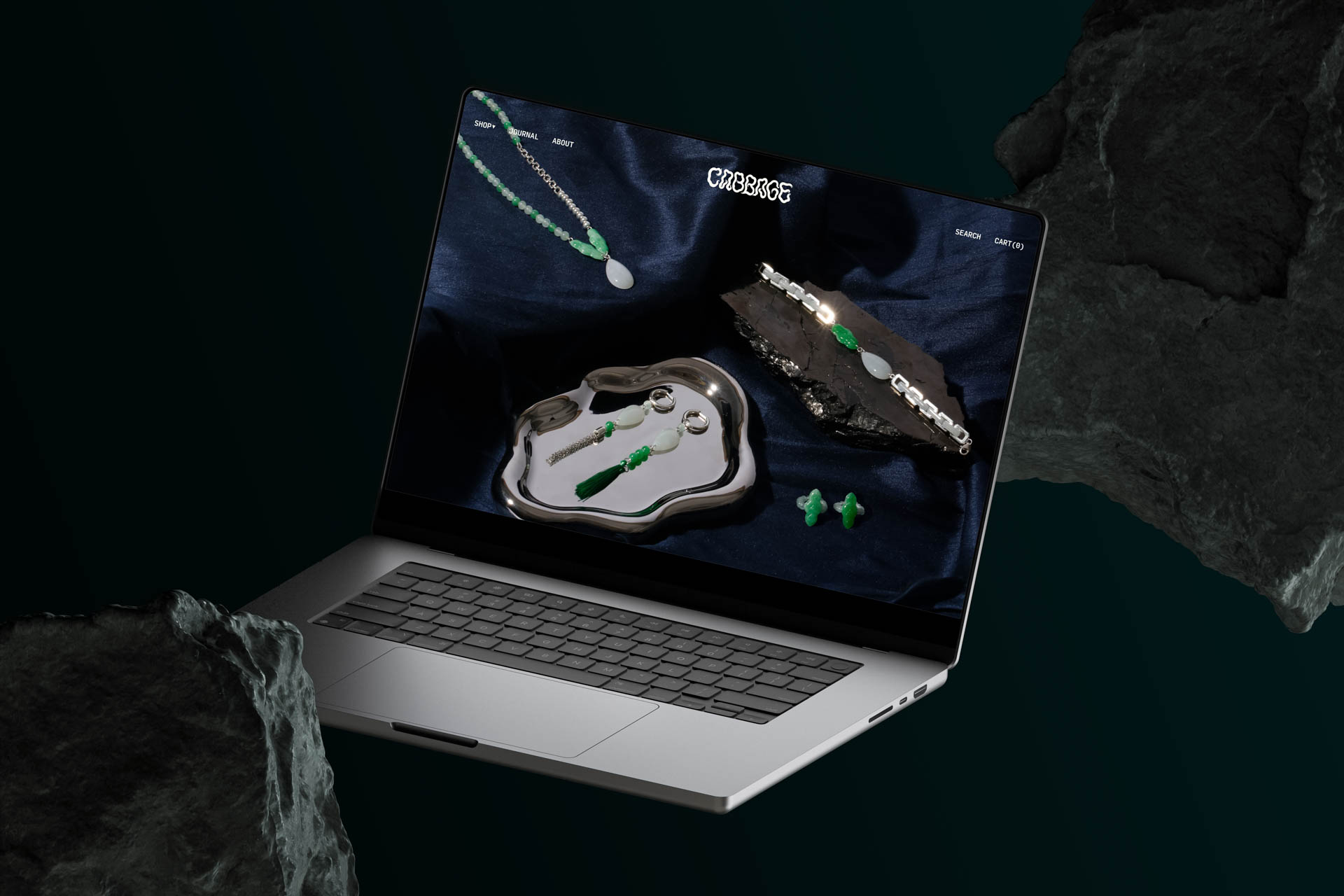 A silver laptop is open and displaying a dark blue screen showing jewelry items: a pendant necklace, a pendant in a bowl-shaped case, and a pair of earrings. The laptop is positioned against a dark green background with rock-like shapes on either side. The screen also displays “CYBERKIDS” at the top and “SEARCH BY CATEGORY” on the right.