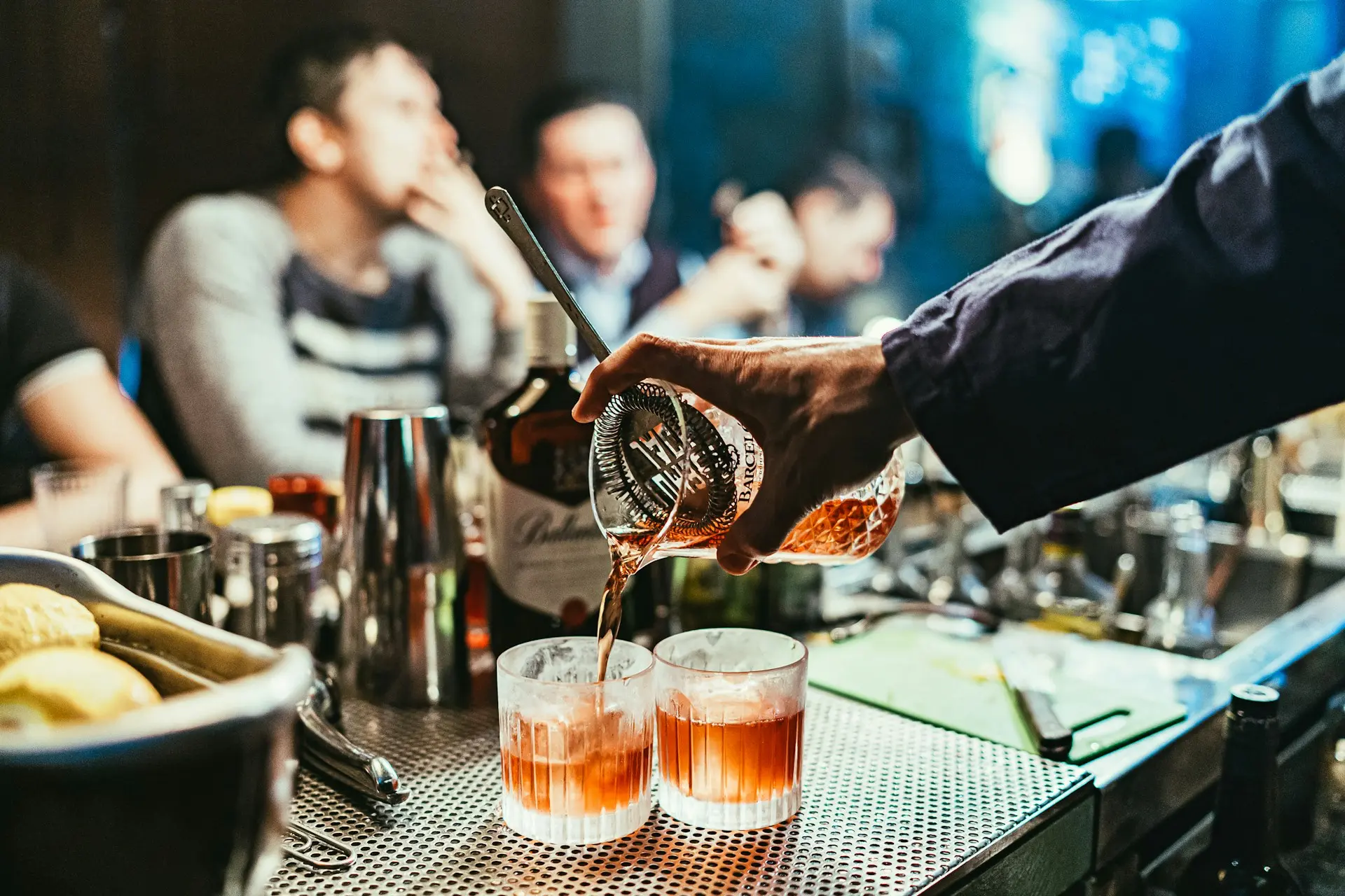 Bartender pouring a cocktail through a strainer into two glasses at a busy bar counter with guests in the background.