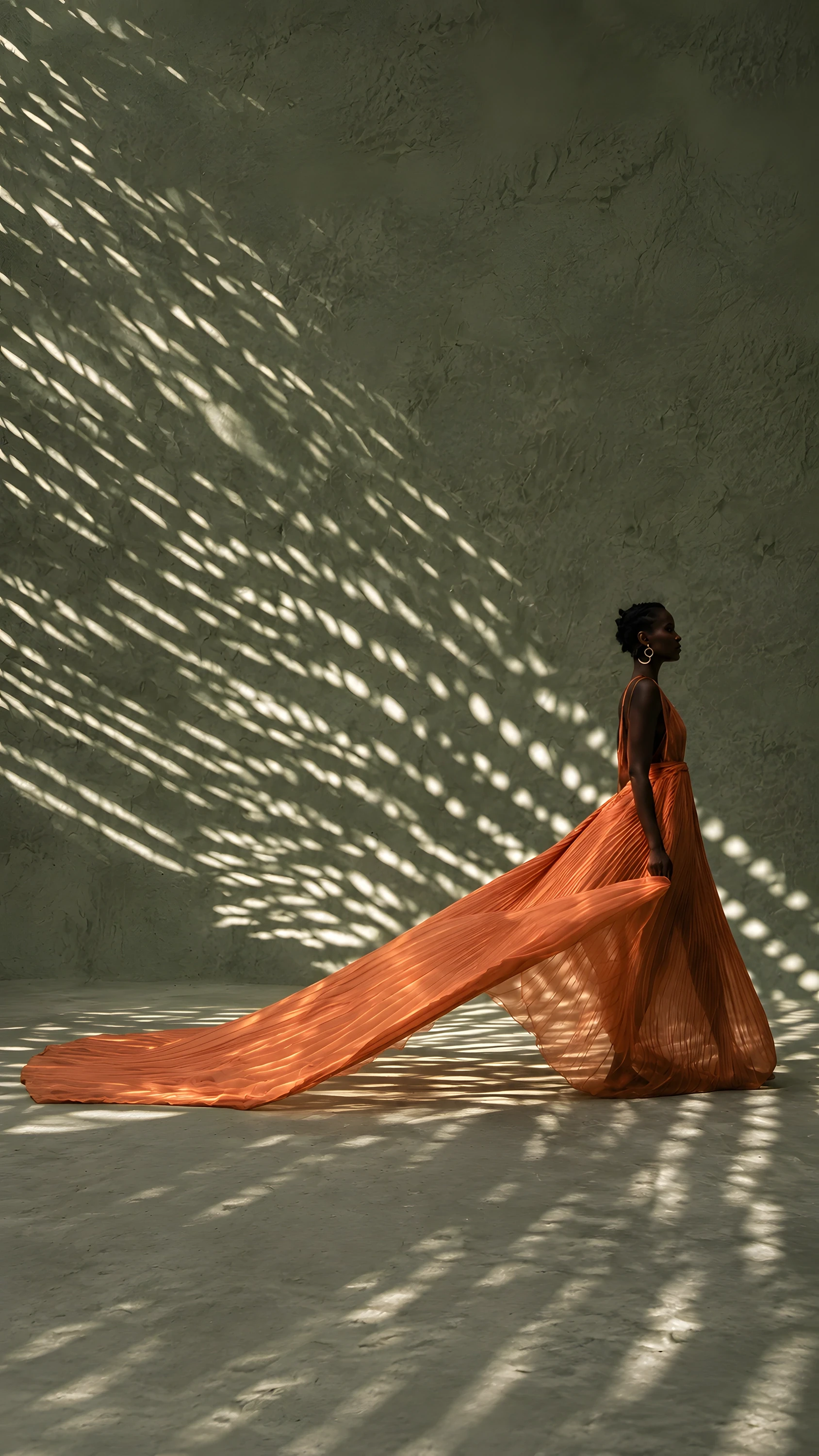 	Woman in a flowing terracotta gown standing against a sage wall patterned with dappled light