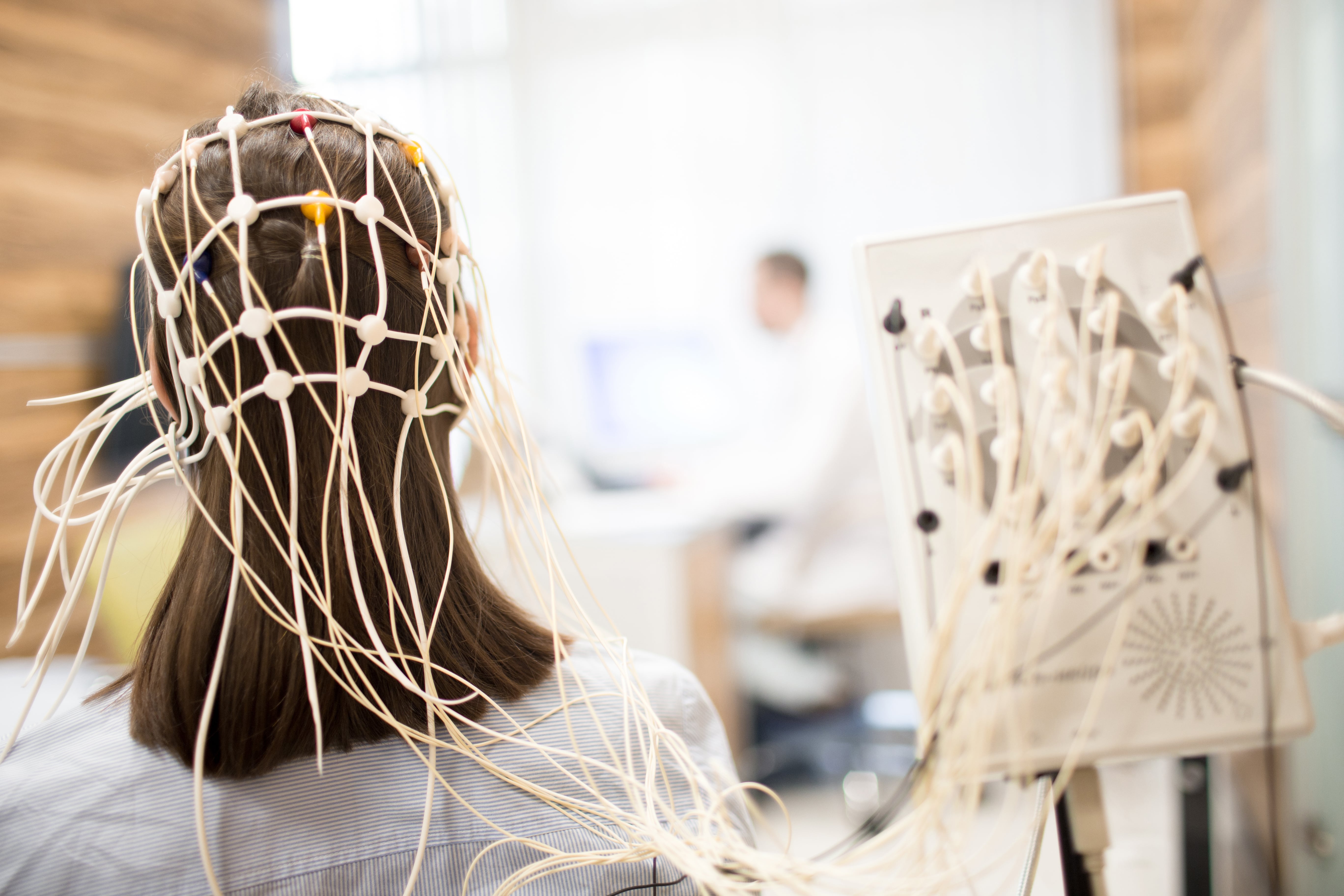 Back view of a female patient wearing an electroencephalography (EEG) cap with electrodes, connected by wires to a machine for a brain scan in a modern medical laboratory.