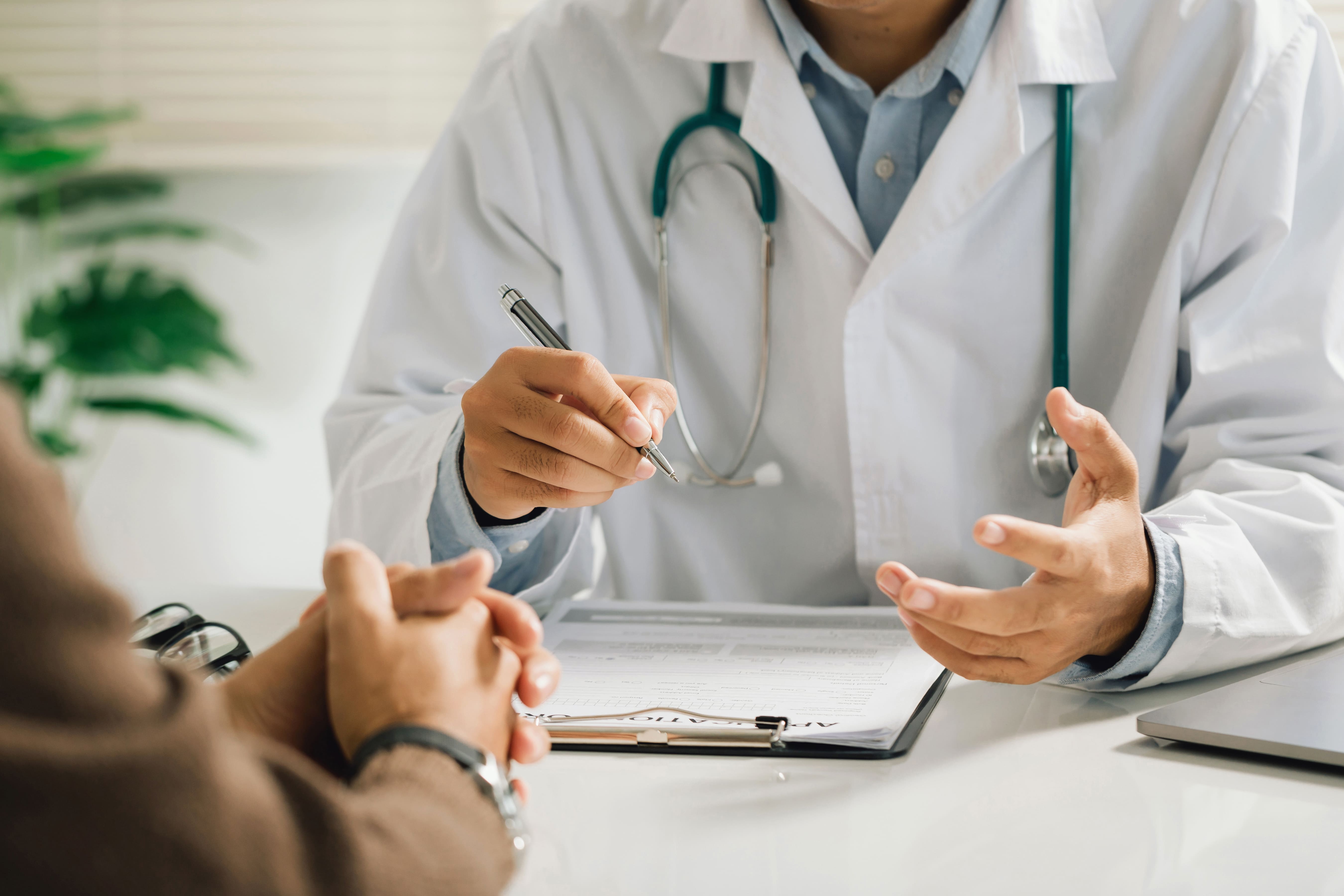 A doctor in a white coat discusses a patient's chart on a tablet across a desk, while the patient listens attentively, highlighting a medical consultation moment.