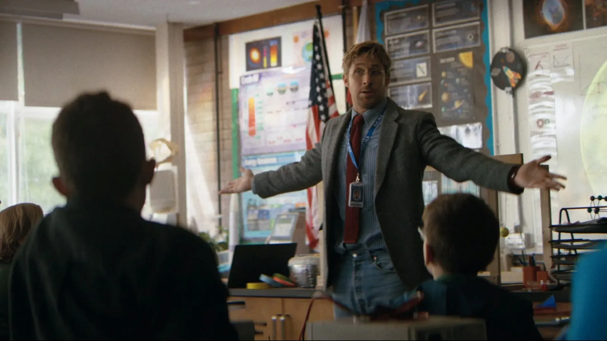 A teacher in a science classroom gestures energetically with arms open, engaging students, with educational posters, an American flag, and scientific models visible in the background, creating a lively learning environment.