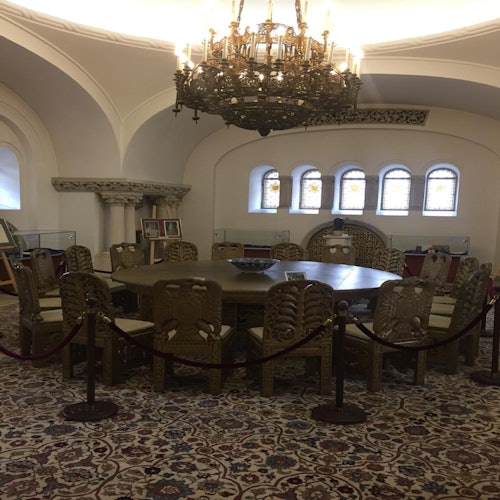 装饰华丽的会议室，配有圆桌和雕刻精美的木椅，周围是彩色玻璃窗，顶部是一盏大型枝形吊灯。