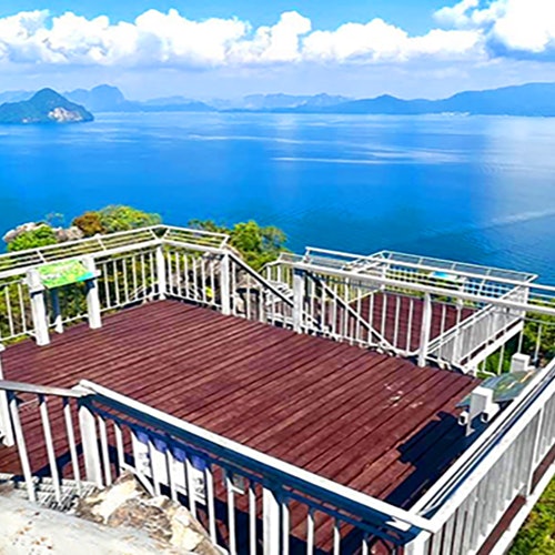 Isang nakaangat na platapormang kahoy na may mga rehas ang sumusulyap sa isang tanawin ng asul na tubig at mga bundok sa ilalim ng bahagyang maulap na kalangitan.