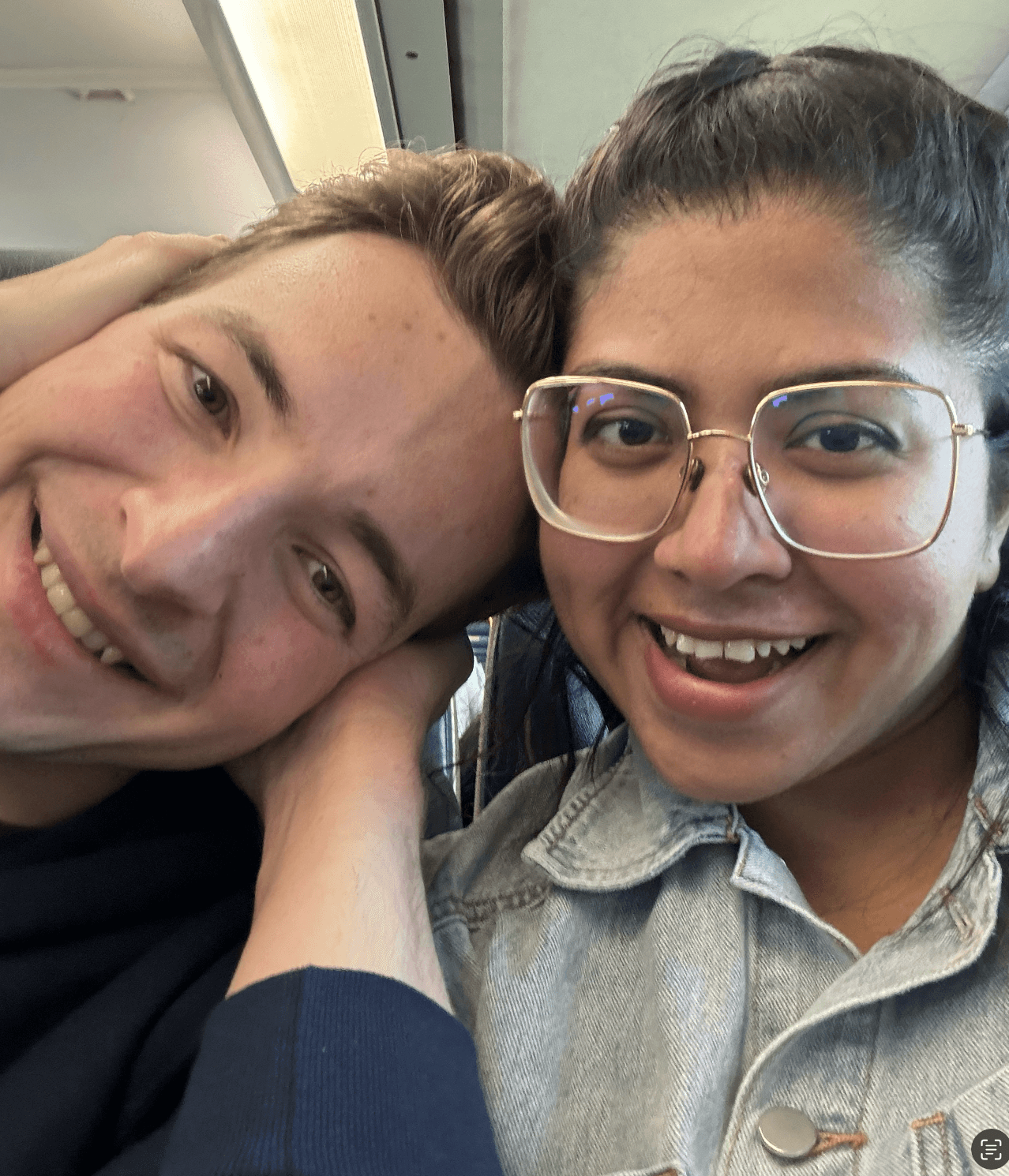 A couple stands on a sandy hill at sunset, facing away, with arms raised in a relaxed "hang loose" gesture. The scene conveys tranquility and joy.