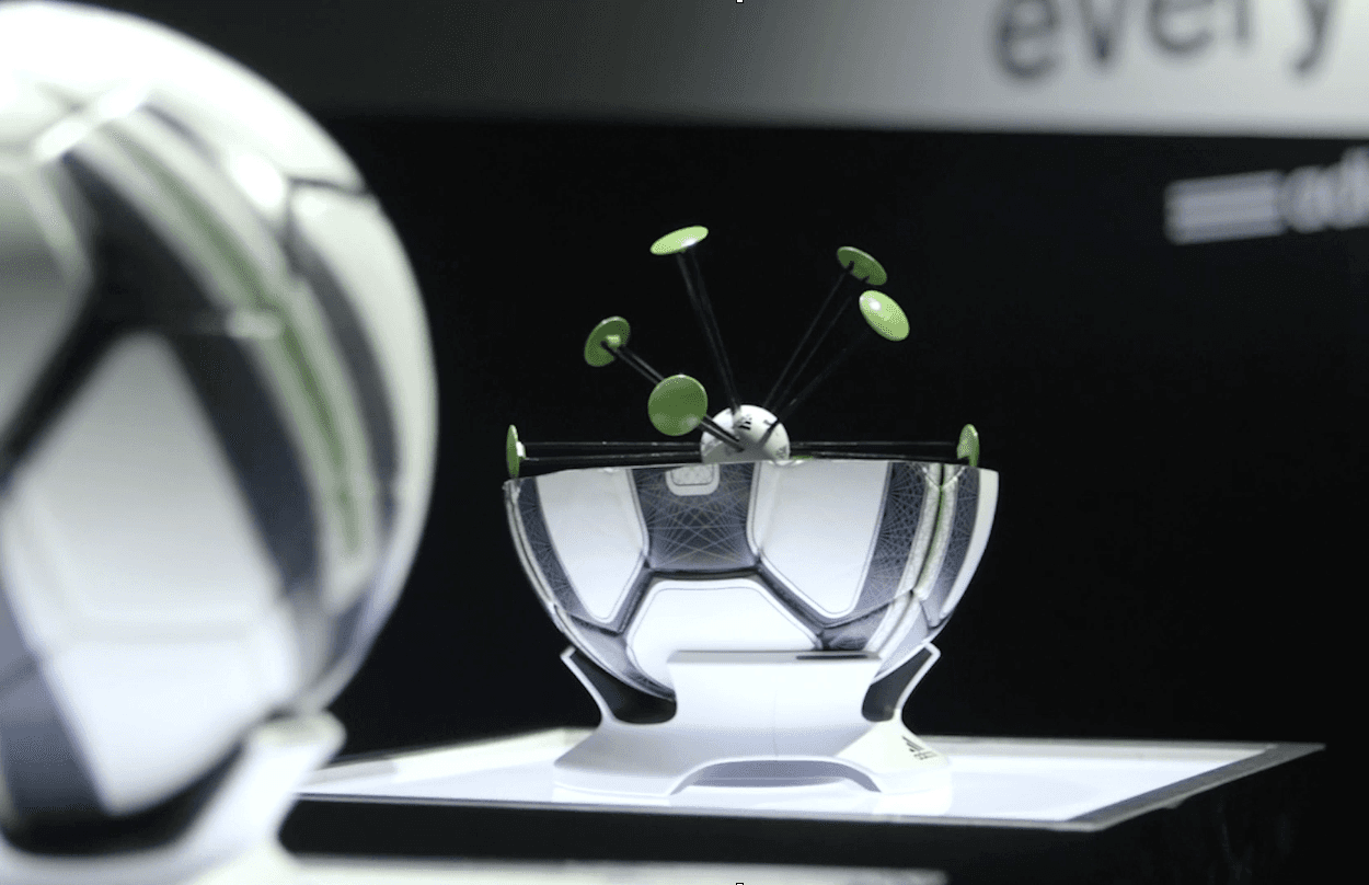 A futuristic soccer ball display features a metallic design with green accents and rotating elements inside a clear, stylized shell, set on an illuminated platform against a dark background.