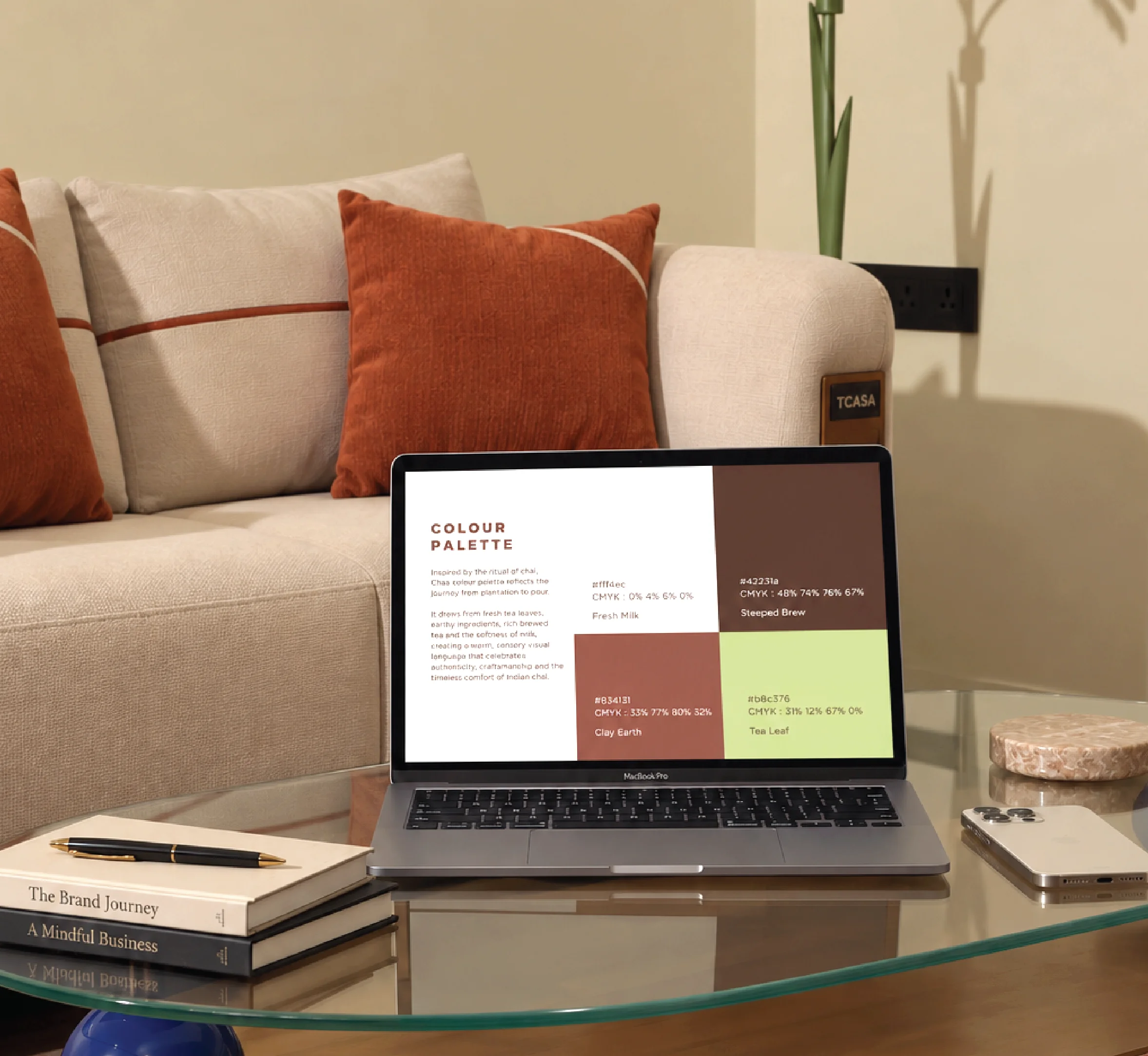 A sleek laptop displaying a color palette sits on a glass coffee table, surrounded by stylish orange and beige cushions, a stack of business books, and minimalist decor in a modern living room setting.