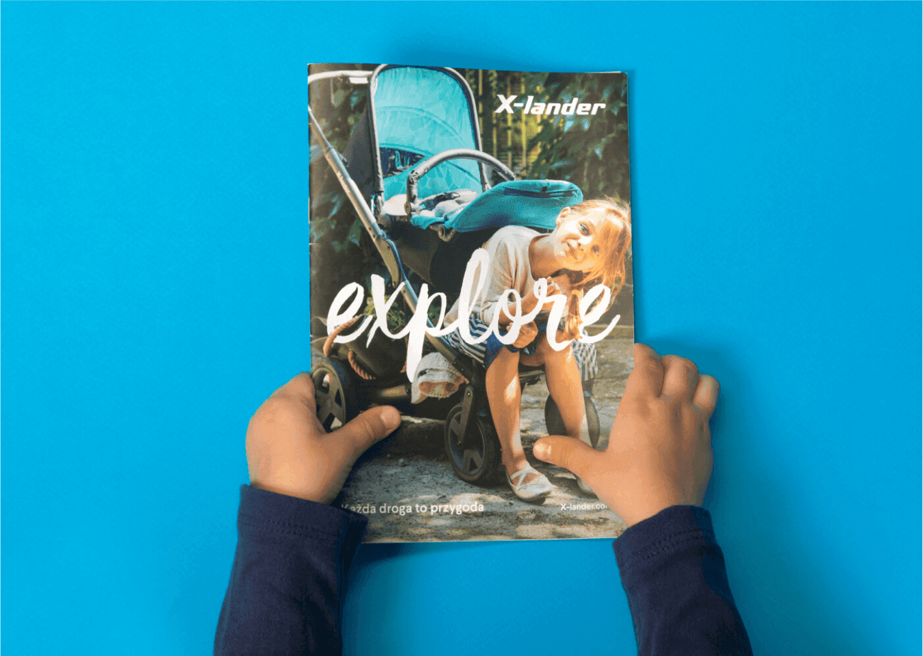 Dziecięce dłonie trzymają katalog z dziewczynką przy turkusowym wózku, napisem „explore” i hasłem „Każda droga to przygoda”.